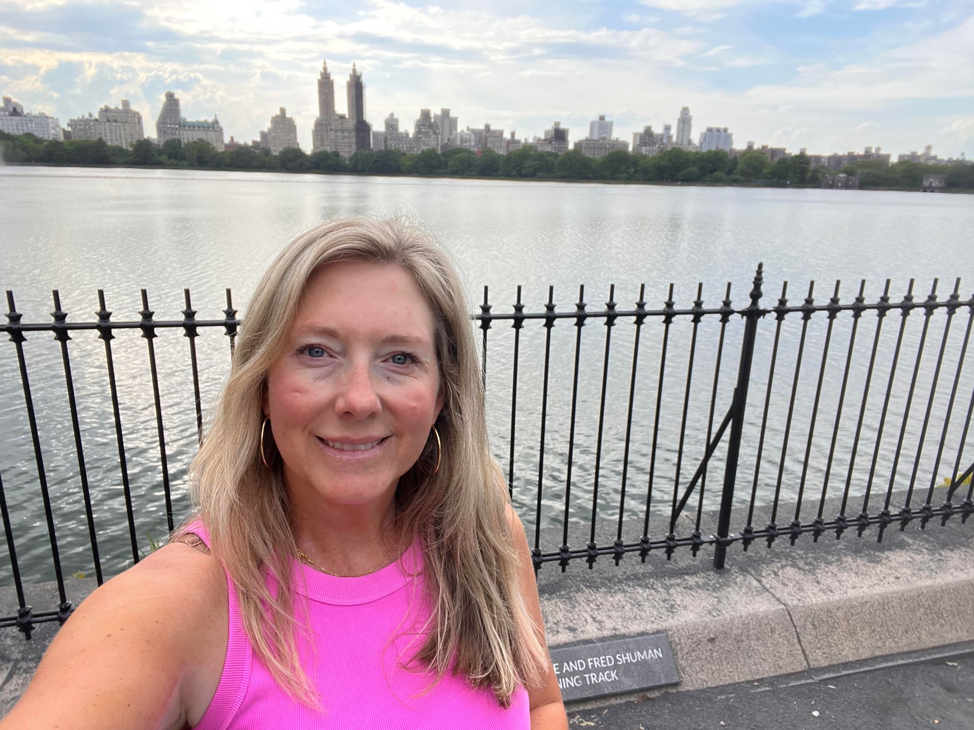 A woman in a bright pink tank top takes a selfie in front of a lake and city skyline in Central Park, New York.
