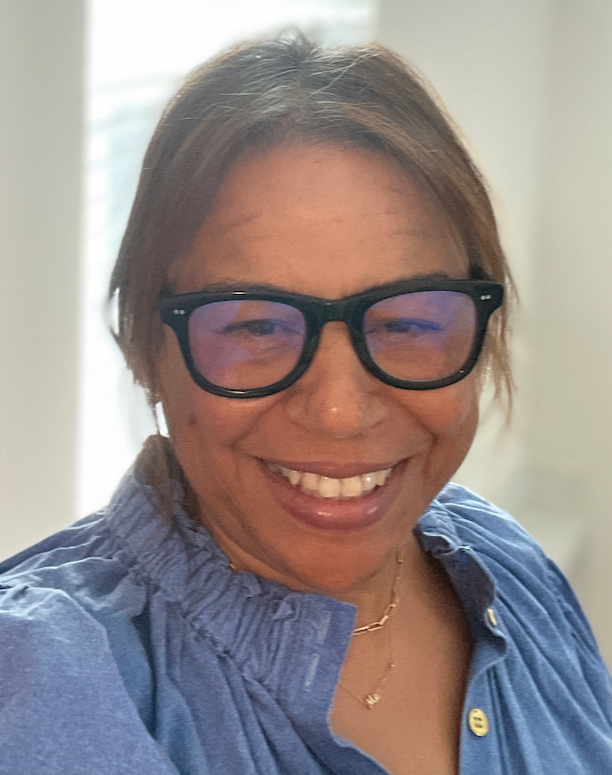 A person with shoulder-length brown hair, wearing black-rimmed glasses and a blue collared shirt, smiling at the camera.
