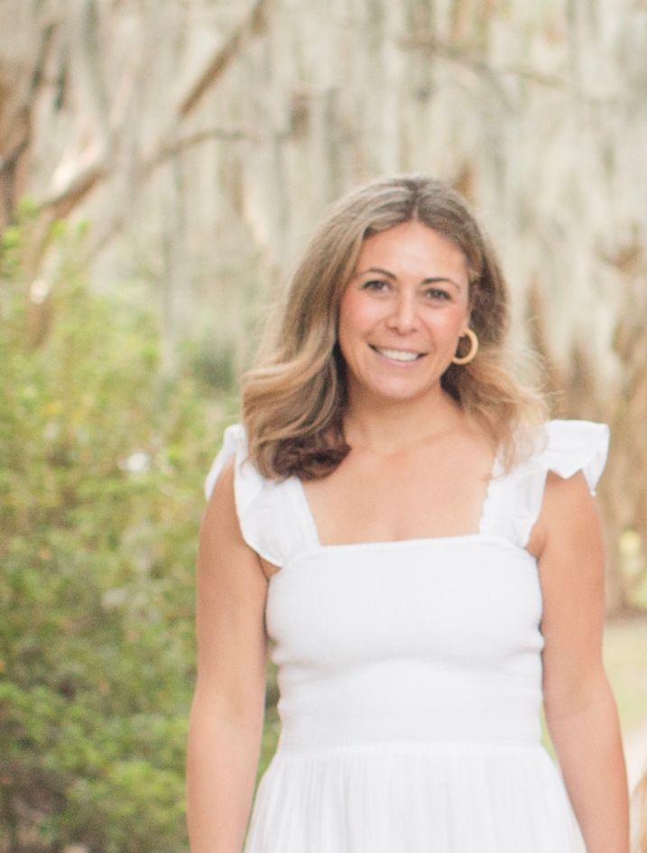 A person with shoulder-length wavy hair wearing a white ruffled-sleeve dress smiles in a garden with hanging tree moss.