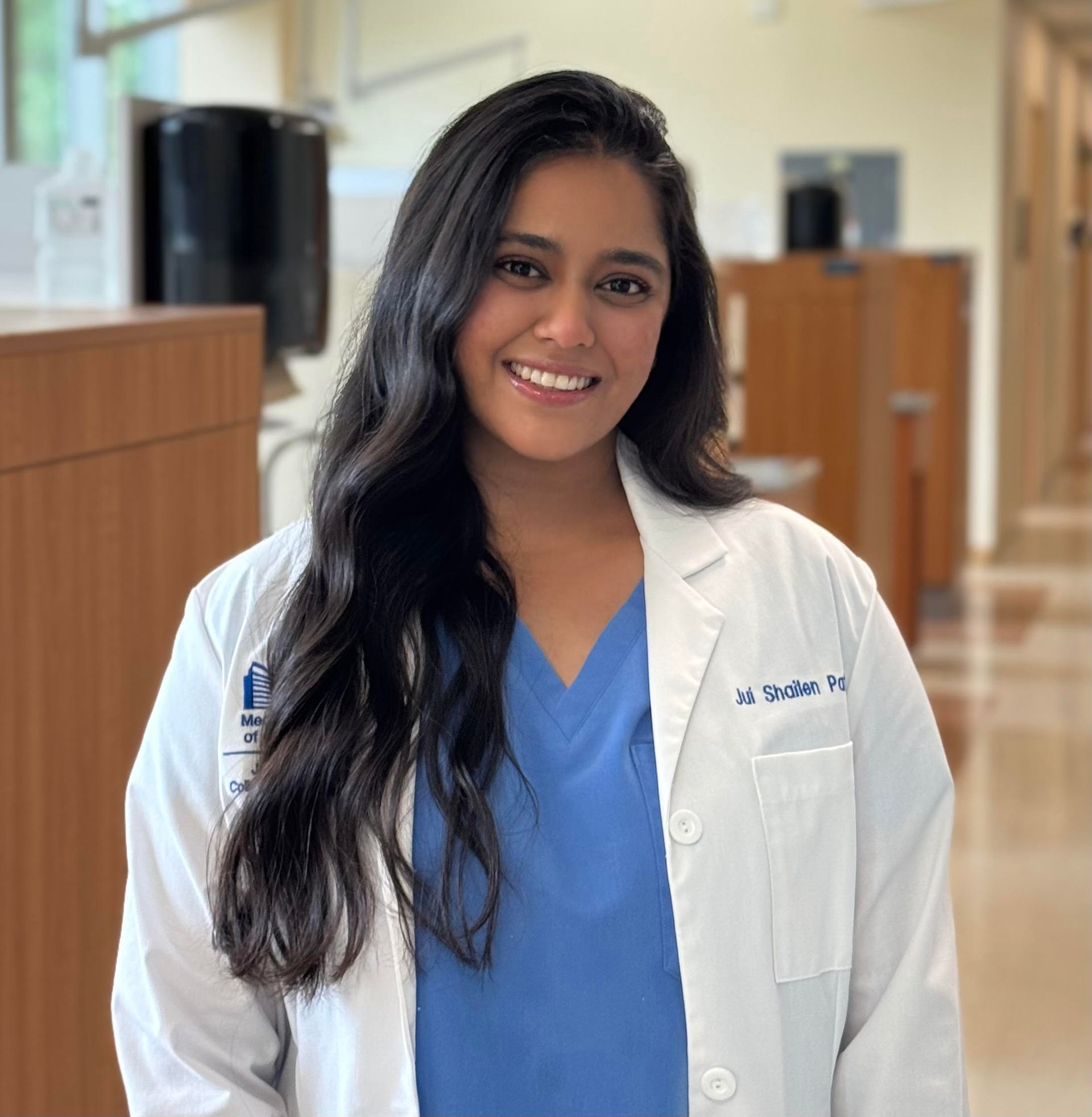 A smiling person wearing a white lab coat over a blue scrub top in a hospital hallway.