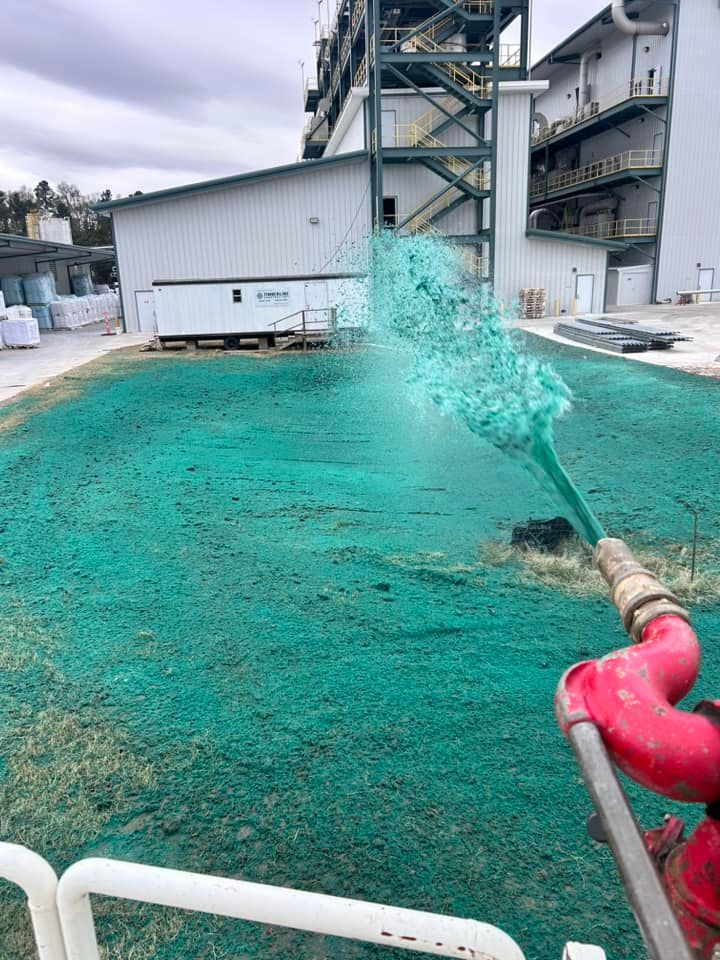 Green hydroseed being sprayed onto bare ground near a large industrial building.