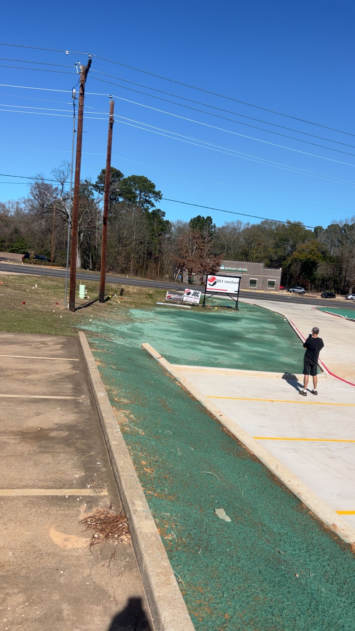 Parking lot with green turf and a person holding a hose on a sunny day. Power lines overhead.