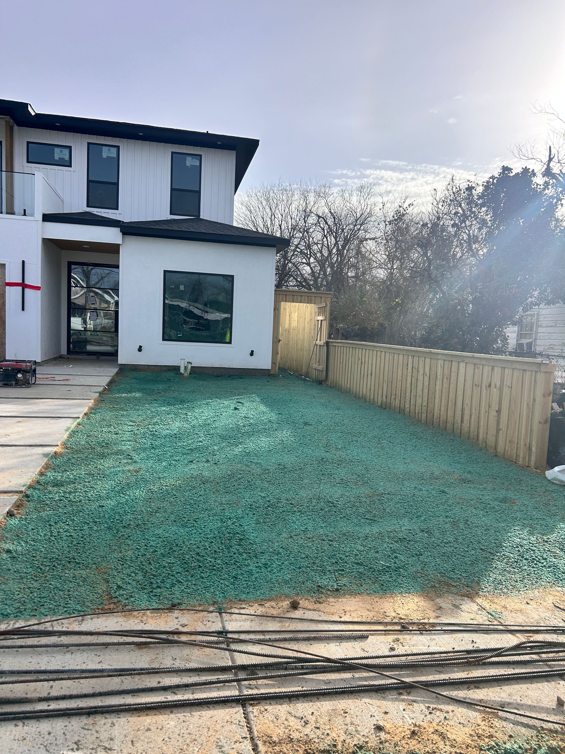 Modern two-story house with newly seeded lawn and wooden fence, next to a sidewalk and street.