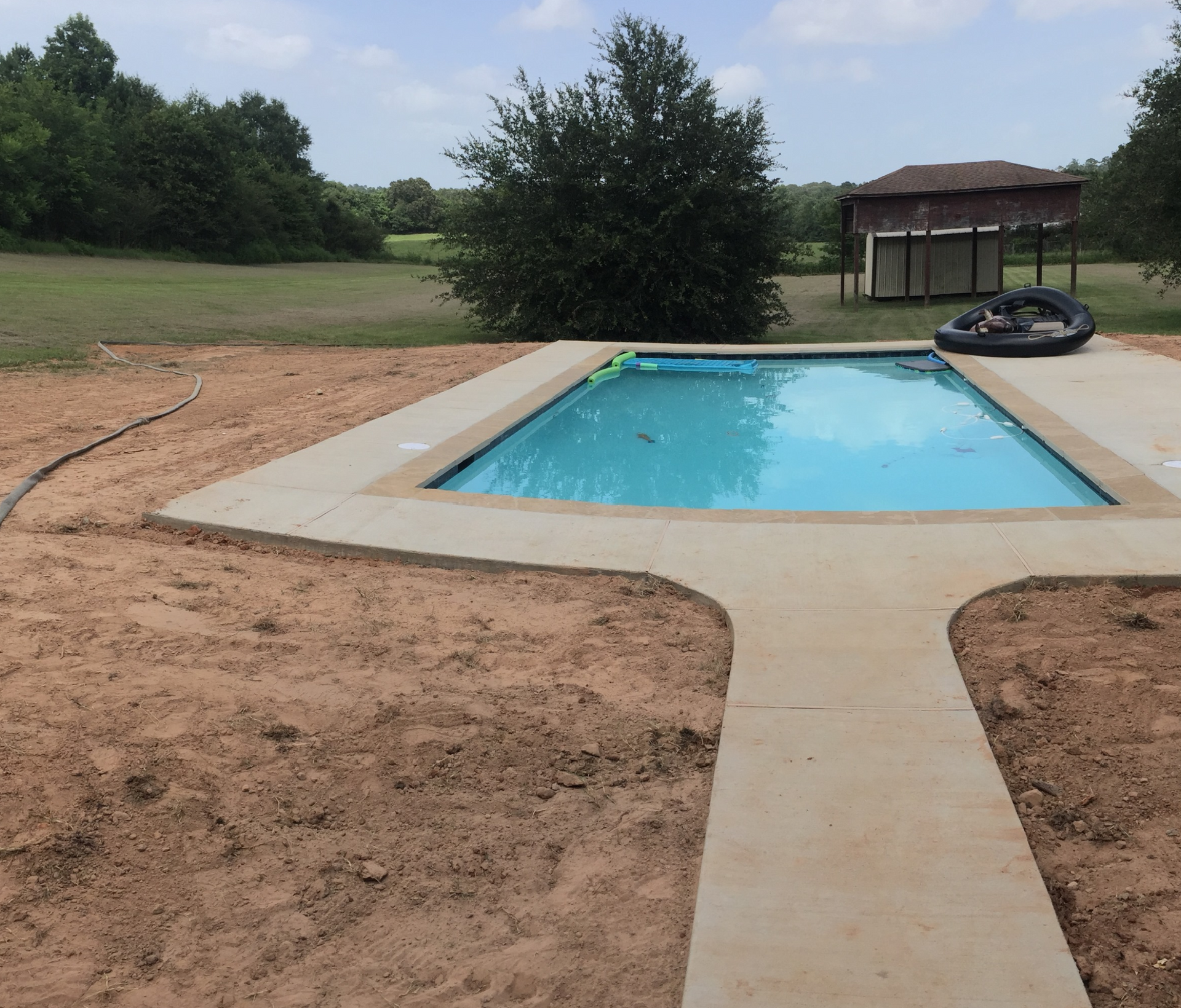 A rectangular swimming pool with concrete decking, surrounded by dirt and grass, and a distant building.