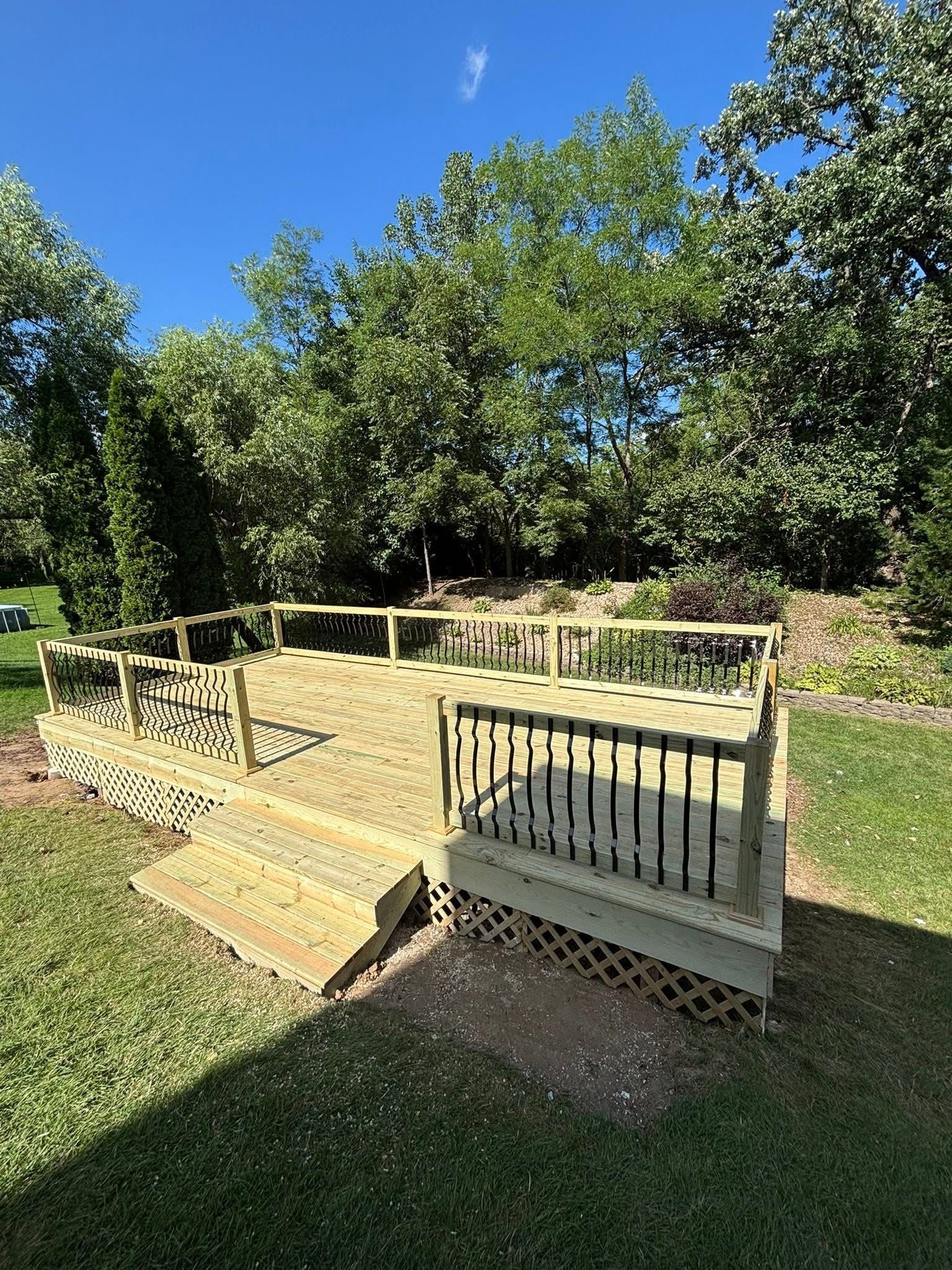 Wooden deck with steps and railing, set on a green lawn, surrounded by trees under a blue sky.
