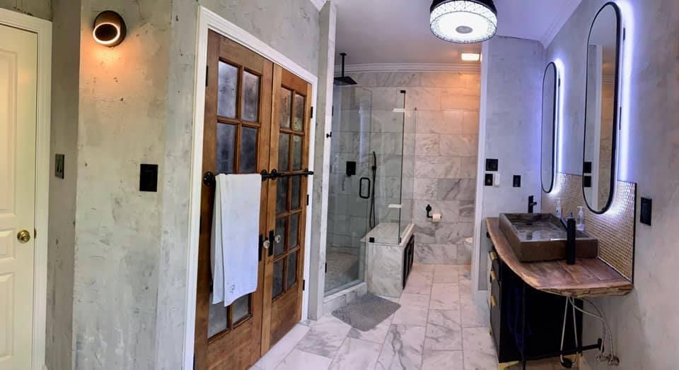 Bathroom with wood door, marble floor, shower, sinks with mirrors, and grey wall.