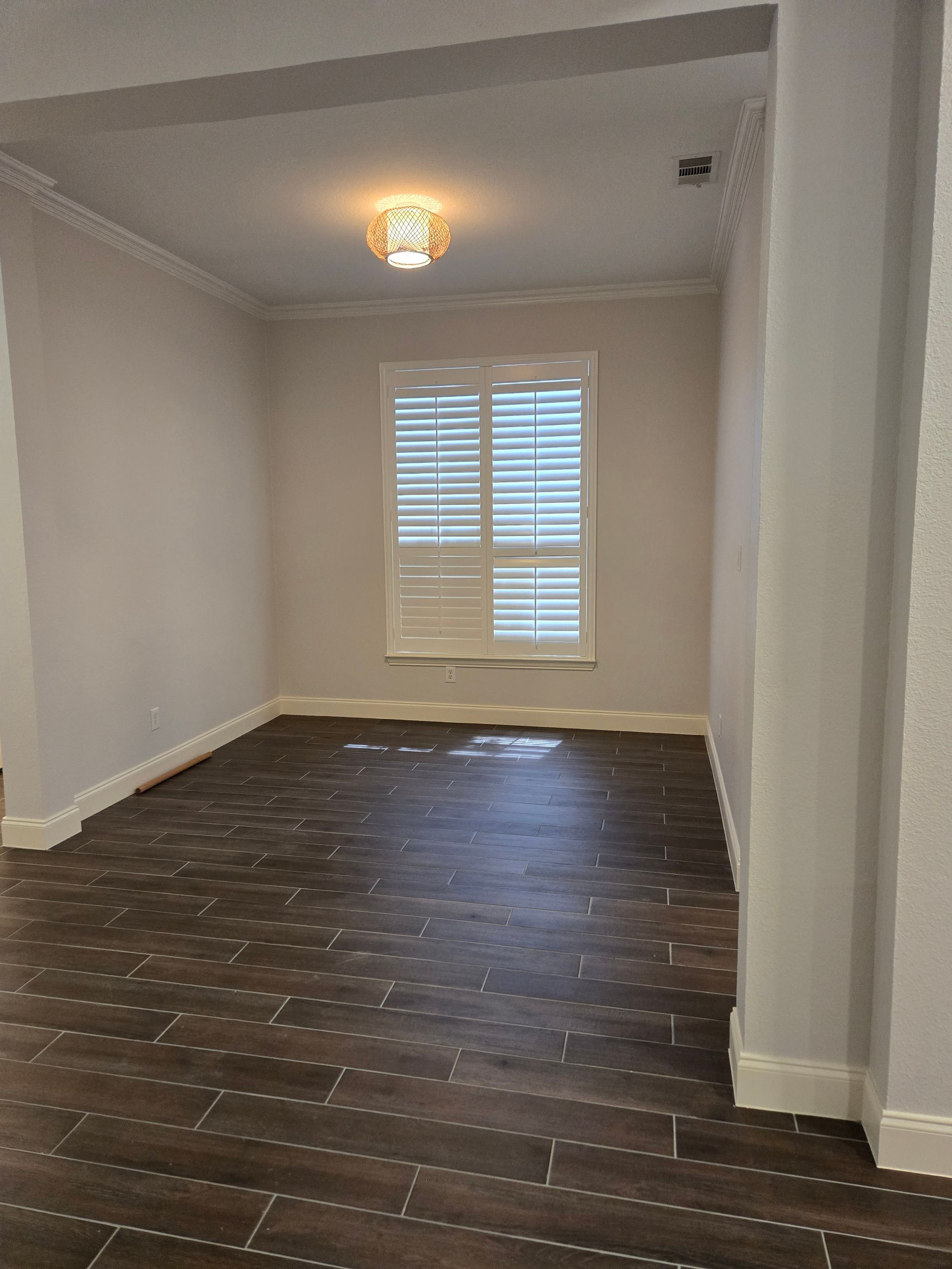 Empty room with wood-look tile floor, white walls and trim, window with shutters, and a decorative light fixture.
