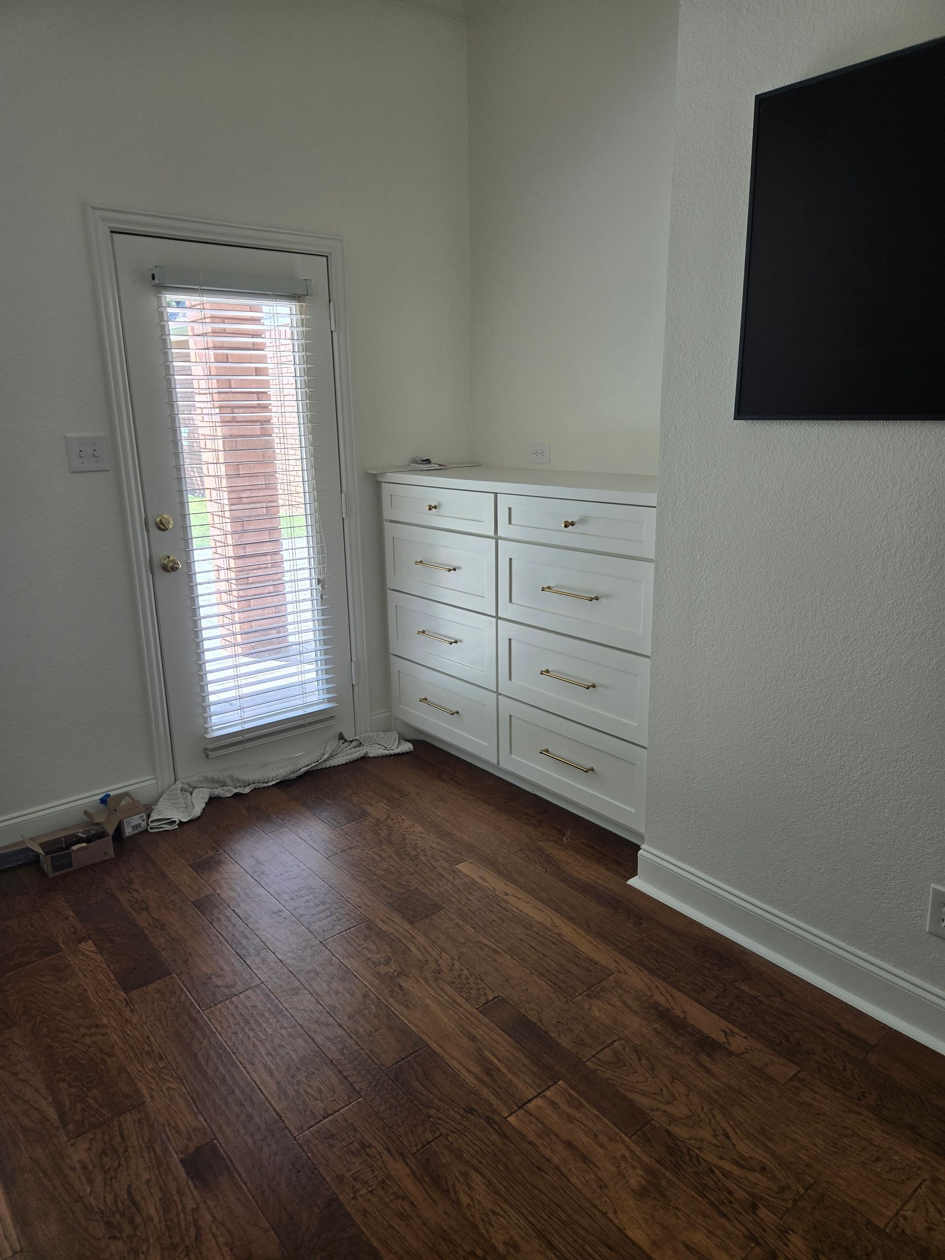 Room with wood floor, white dresser, and glass door to outside. A TV is on the right.