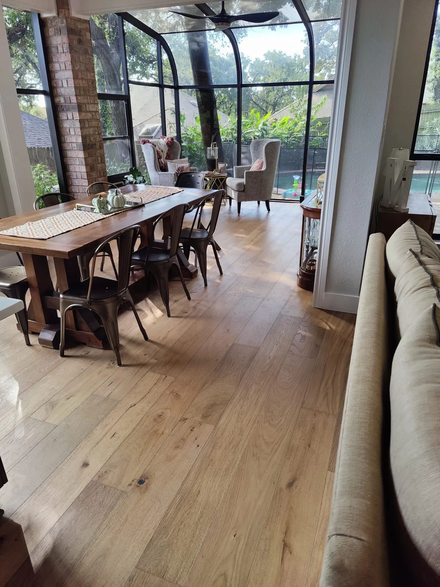 Bright dining room with long wooden table, chairs, and large arched windows overlooking greenery