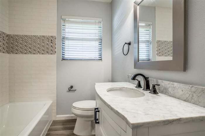 Bathroom with white vanity, marble countertop, bathtub, toilet, and window with blinds.
