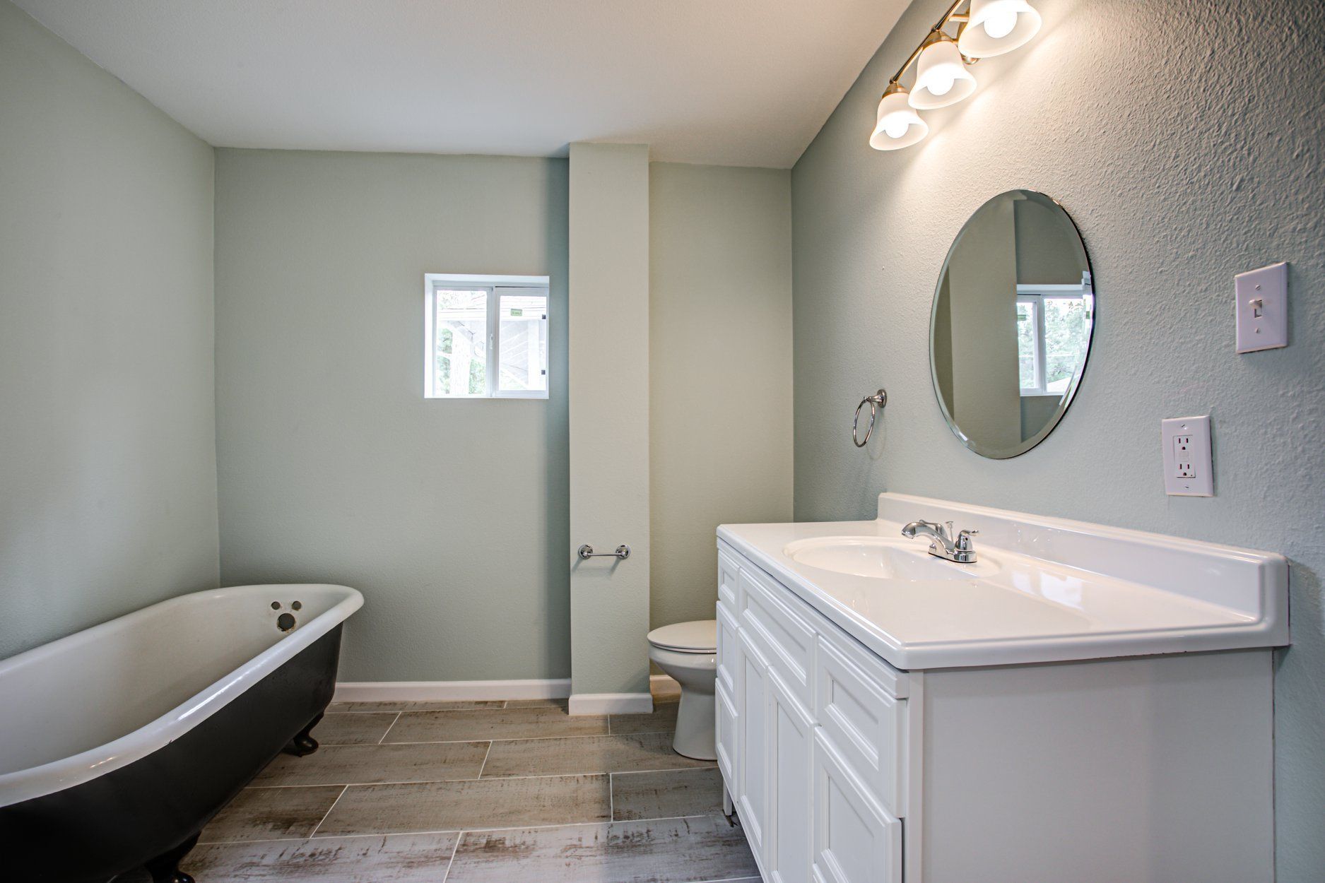 Bathroom with a claw-foot tub, white vanity, and round mirror; pale green walls and gray tiled floor.