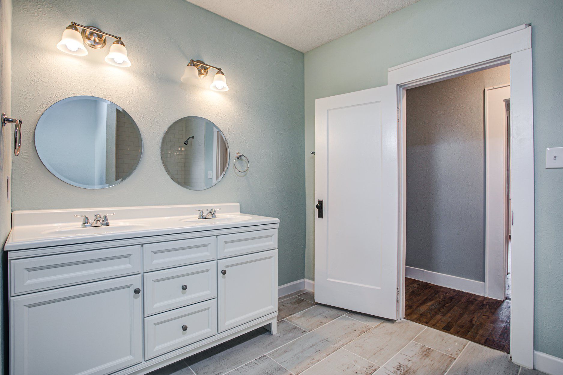 Bathroom with double vanity, round mirrors, and open door. White cabinets and light blue walls.