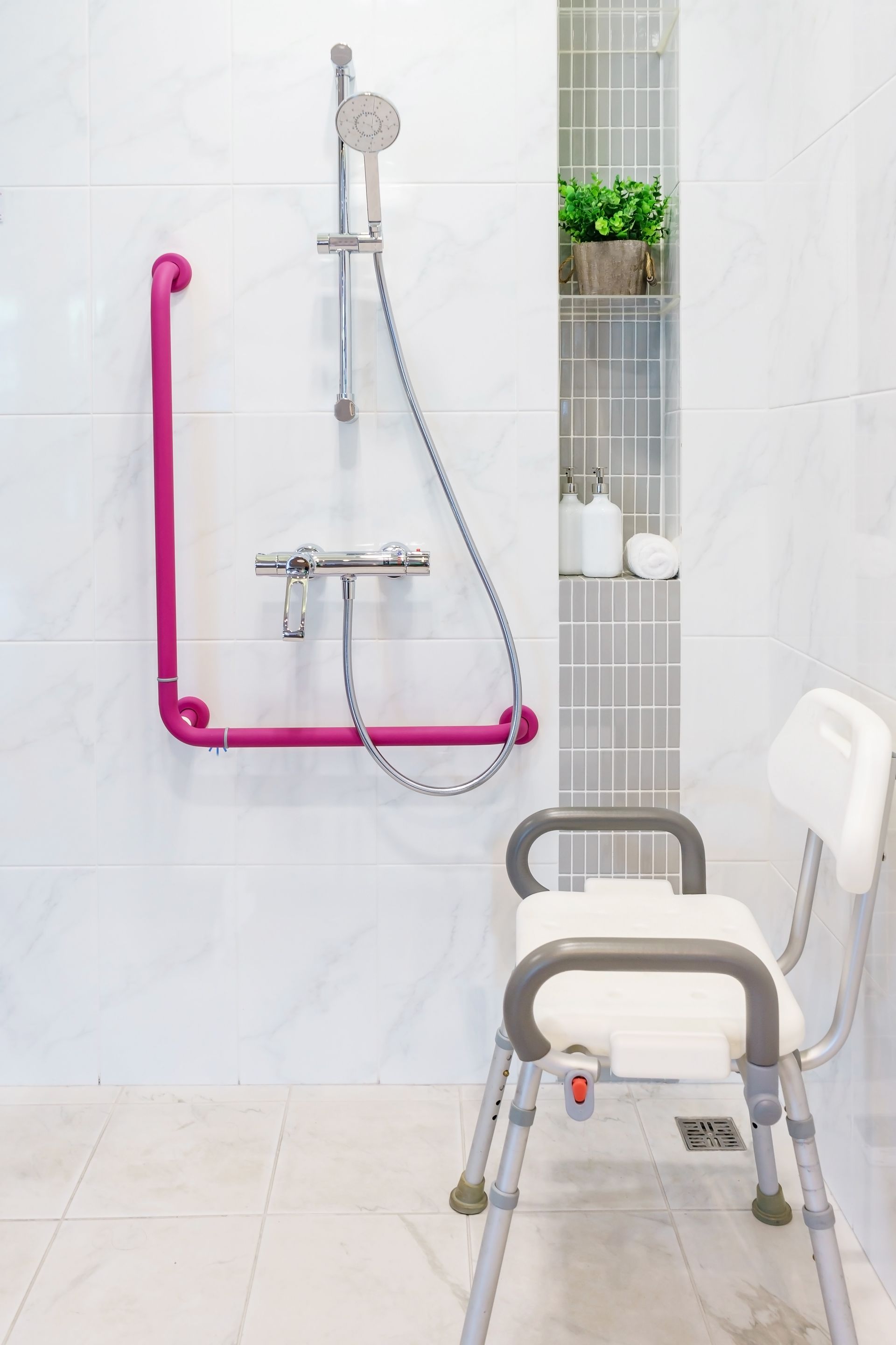 Salle de bain accessible avec siège de douche, barre d'appui rose, flexible de douche et étagère murale avec plantes et articles de toilette.