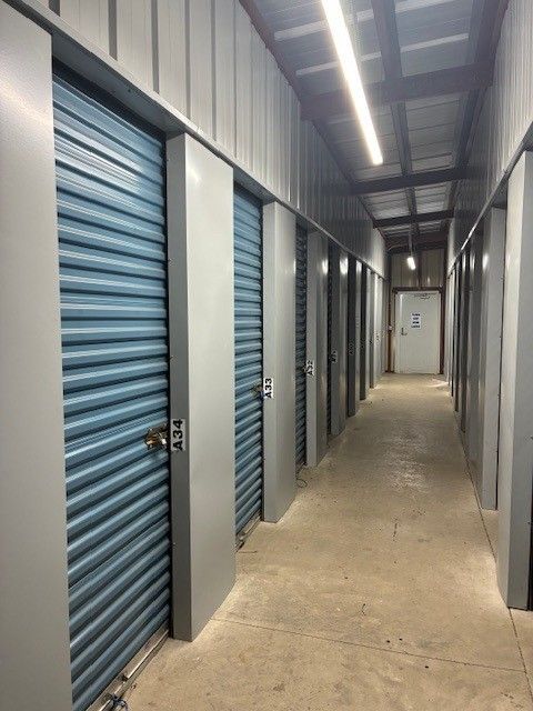 Interior of a storage facility hallway. Blue storage unit doors line the corridor, with a door at the end.