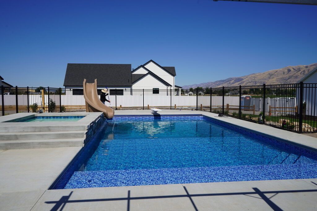 Backyard pool with a slide, hot tub, and house under a blue sky.