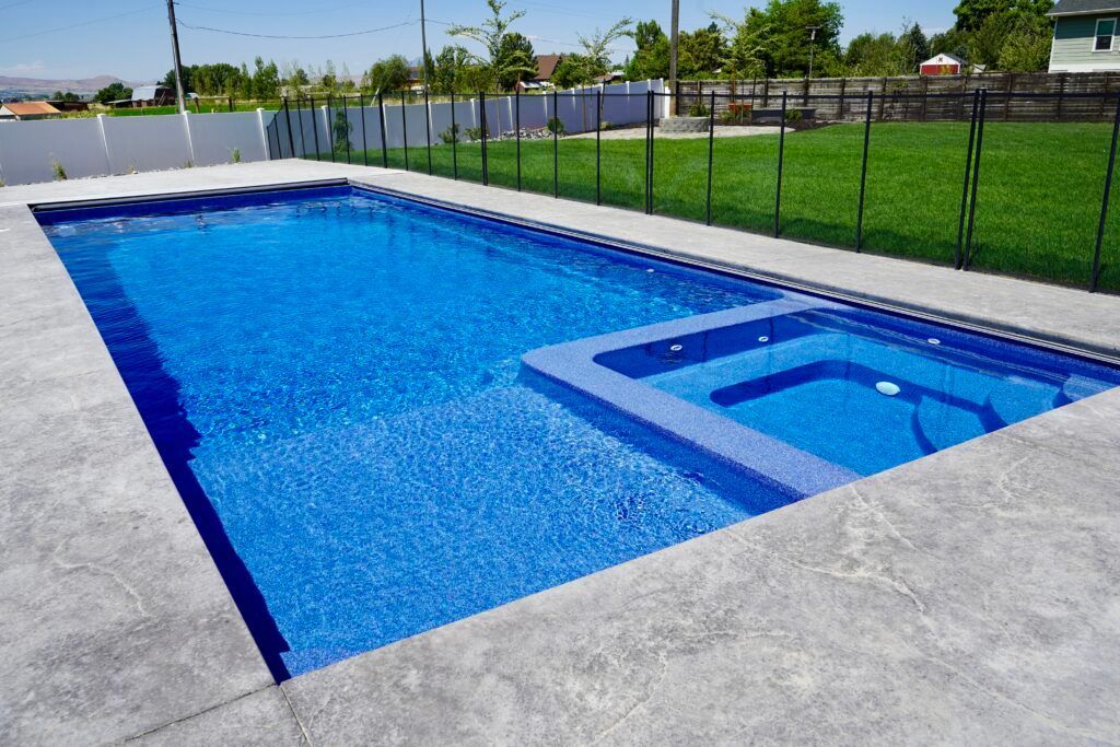 Rectangular blue pool with attached hot tub, set in a backyard with a gray concrete surround and green lawn.