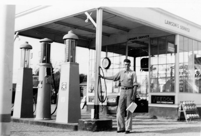 Vintage black-and-white gas station with a man standing beside fuel pumps under a canopy.