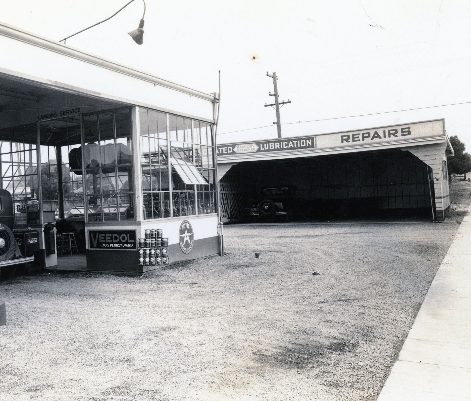Black-and-white view of an auto repair garage with a large open bay and gravel lot.