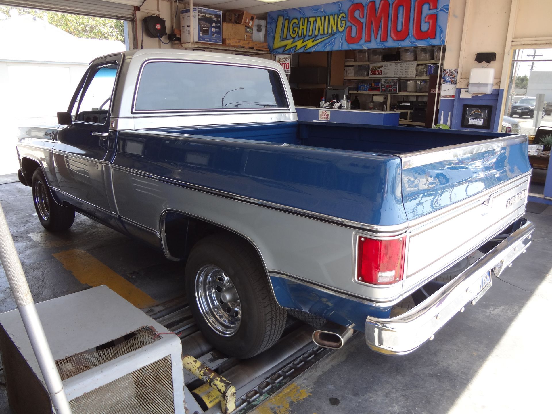 Blue pickup truck parked outside a smog shop, rear three-quarter view