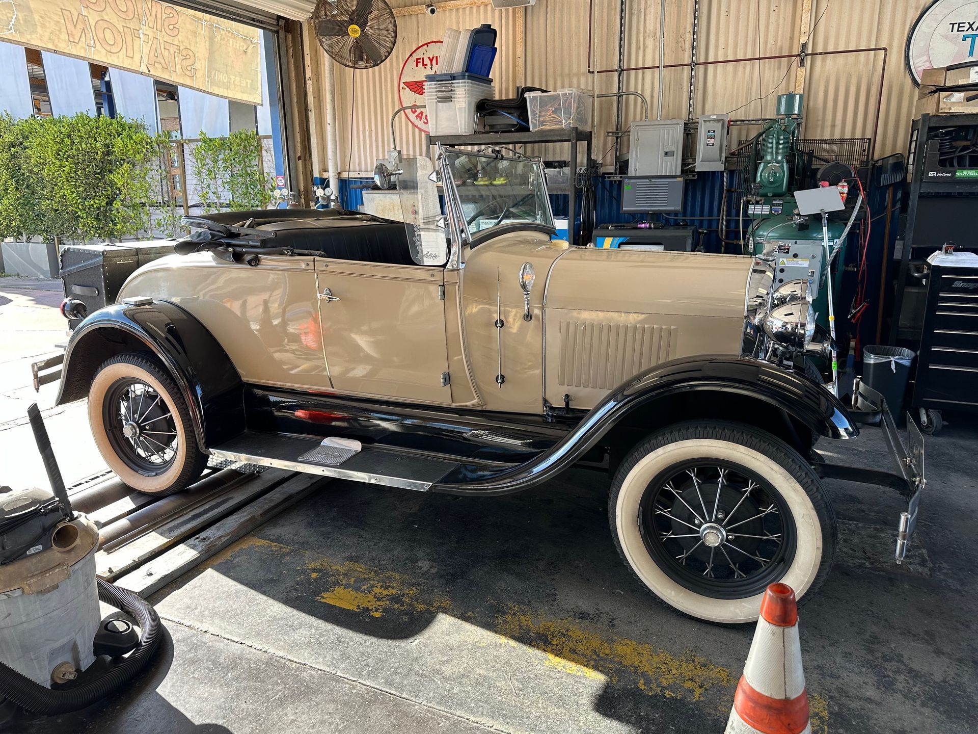 Vintage beige convertible car in a garage, parked beside an orange cone and tools.