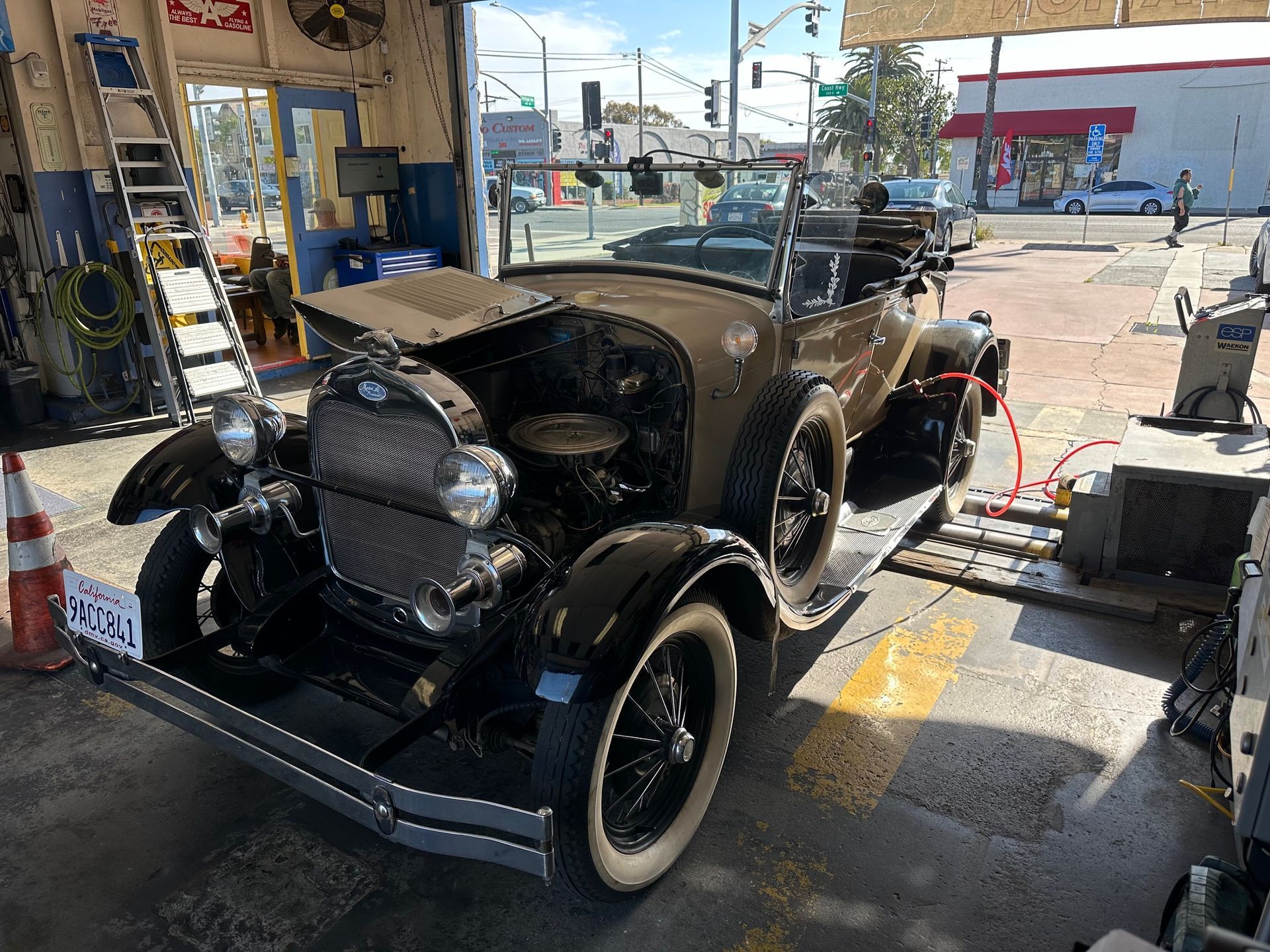Black vintage car at a gas station, with a race number and exposed engine, parked near fueling equipment.