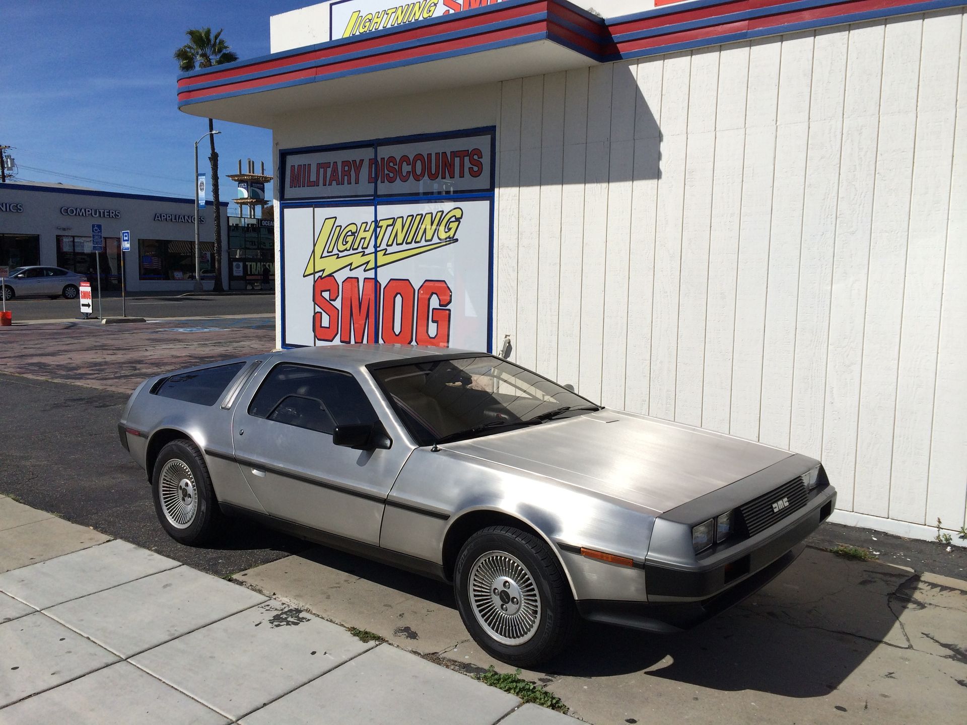 Silver sports car parked outside a smog check shop with a large red-and-yellow sign