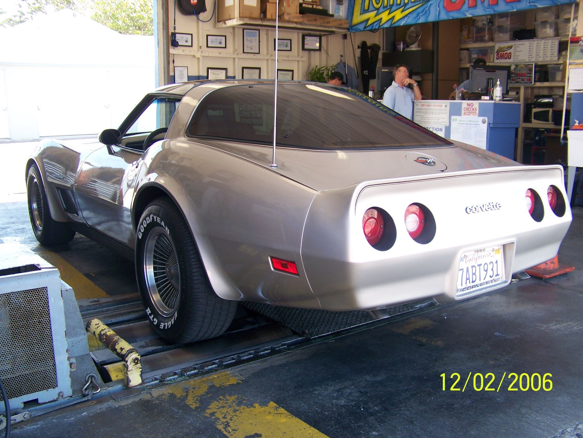 Silver Corvette on a shop lift inside an auto garage, with two men in the background.