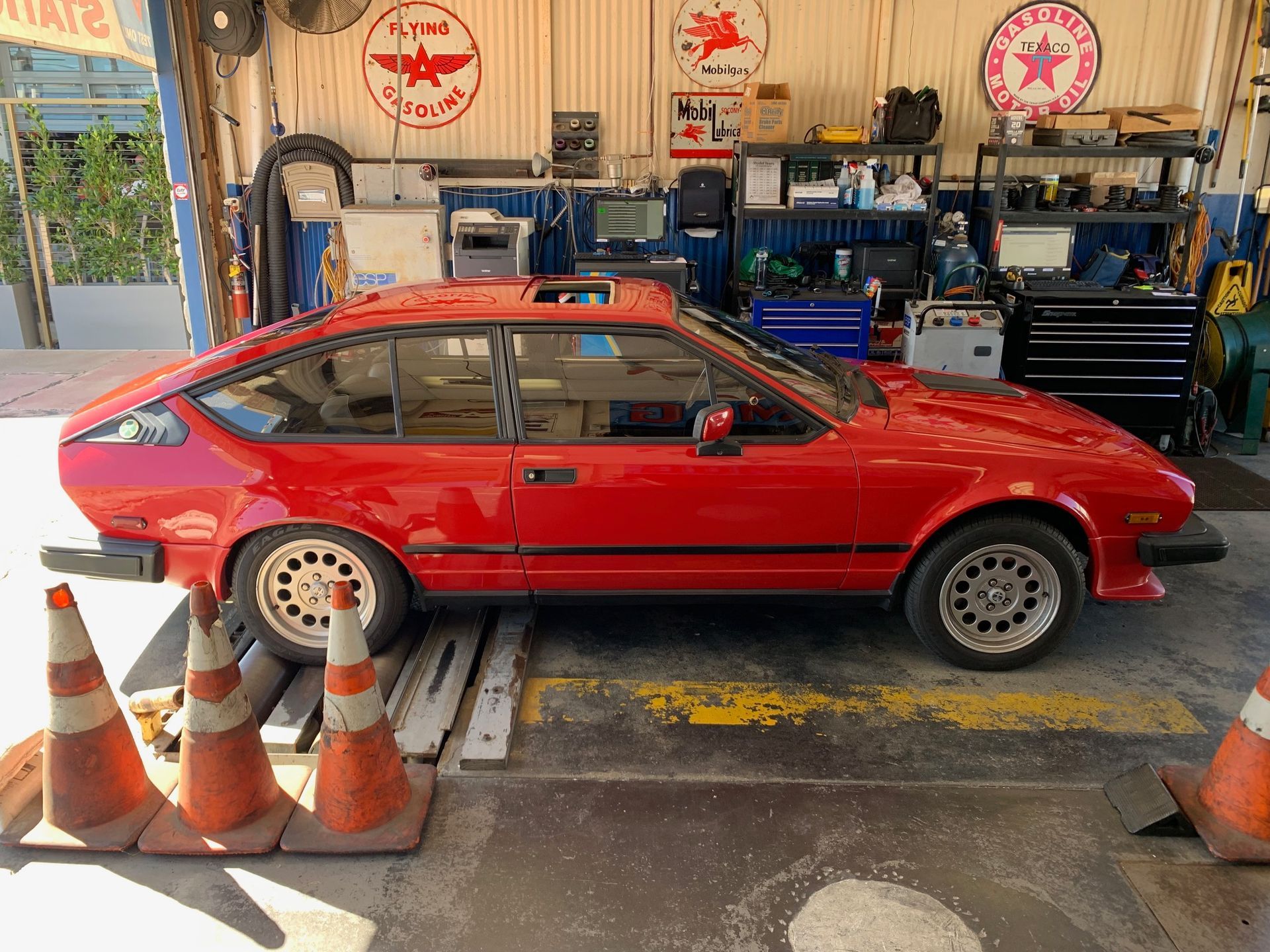 Red sports coupe on a garage lift, with orange cones and shop tools in the background.