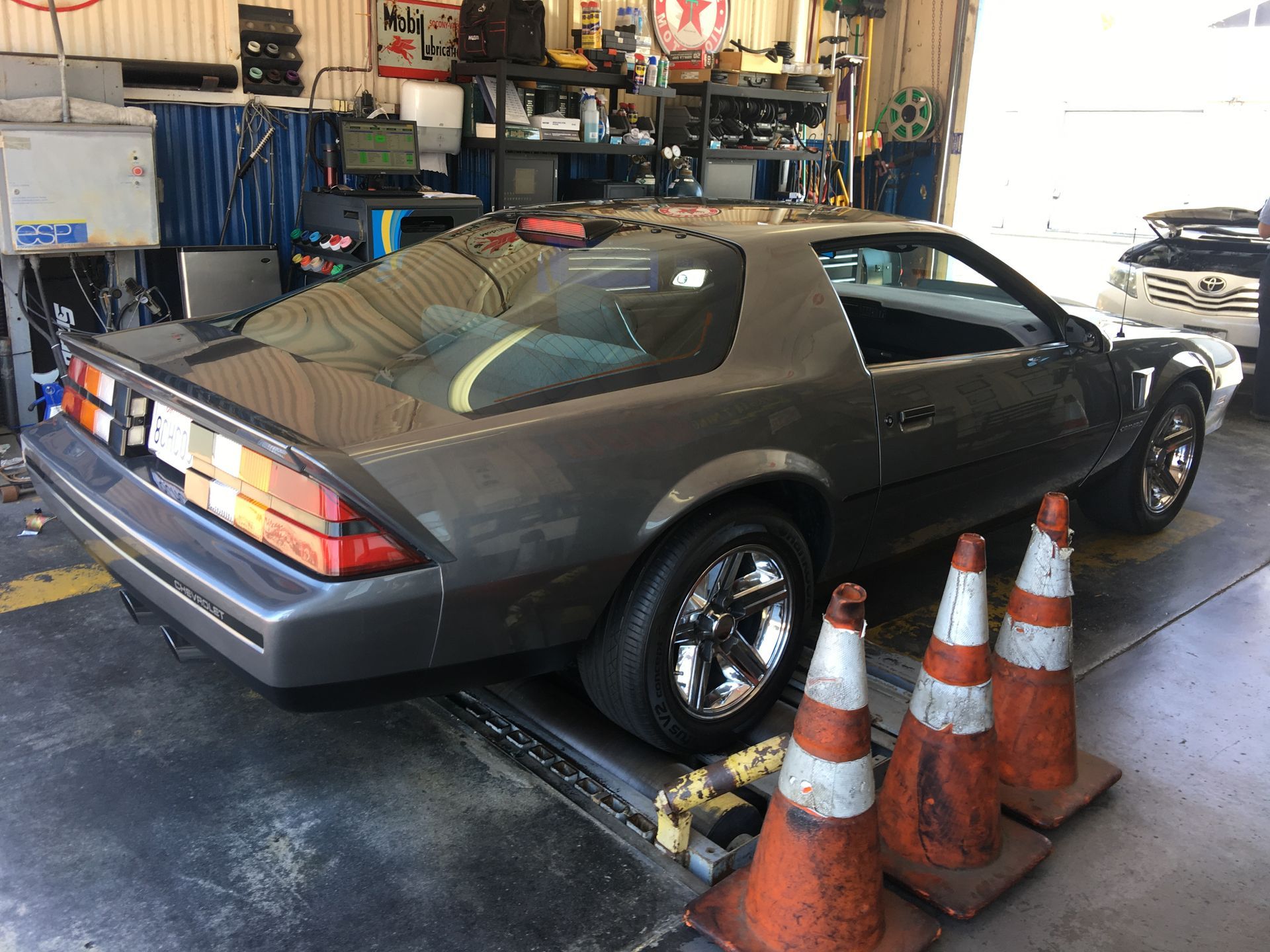 Gray sports car in a garage, rear three-quarter view, with orange cones beside it.