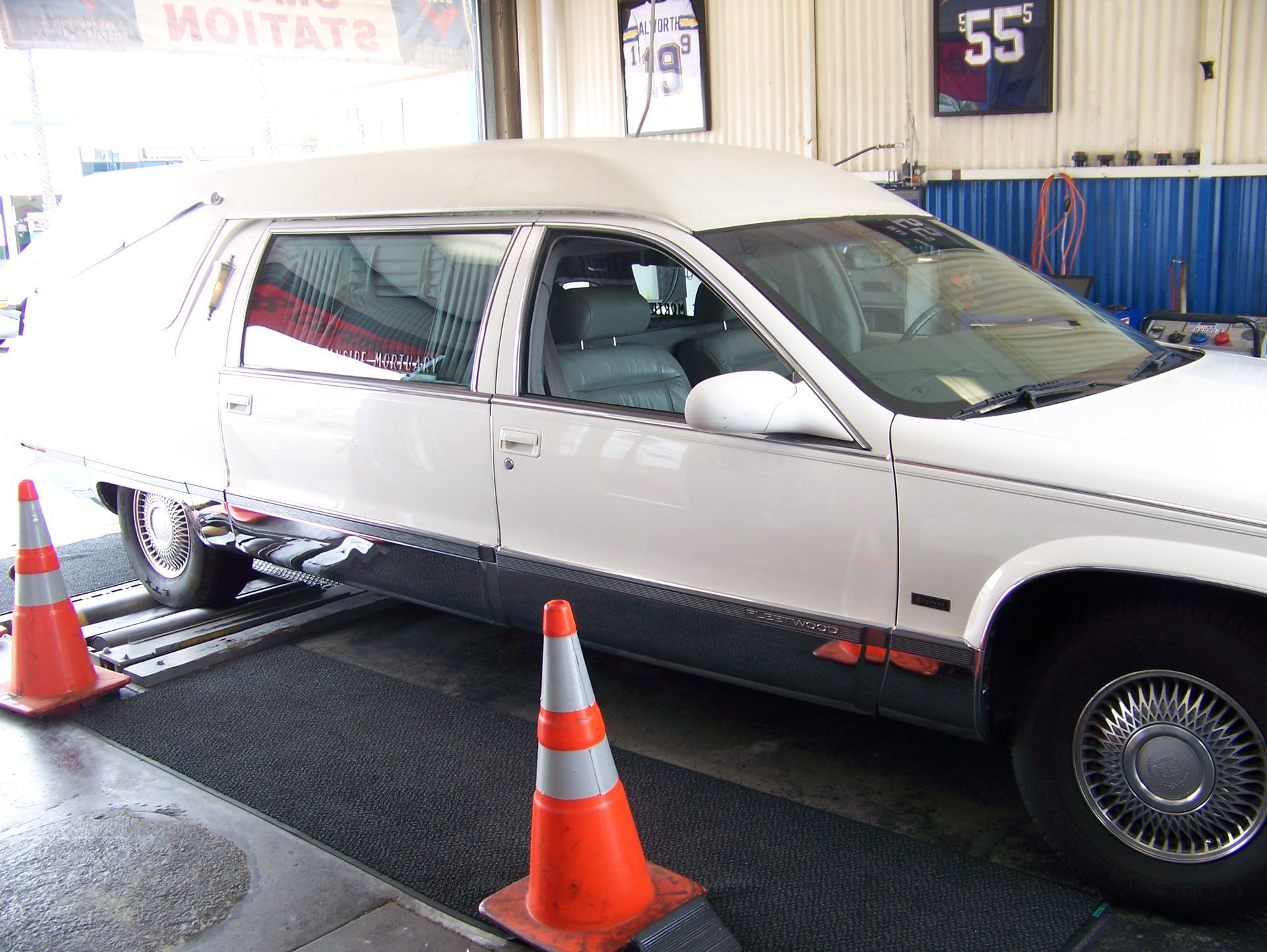 White hearse at a car wash bay, with orange cones and wet floor, being inspected inside a garage.