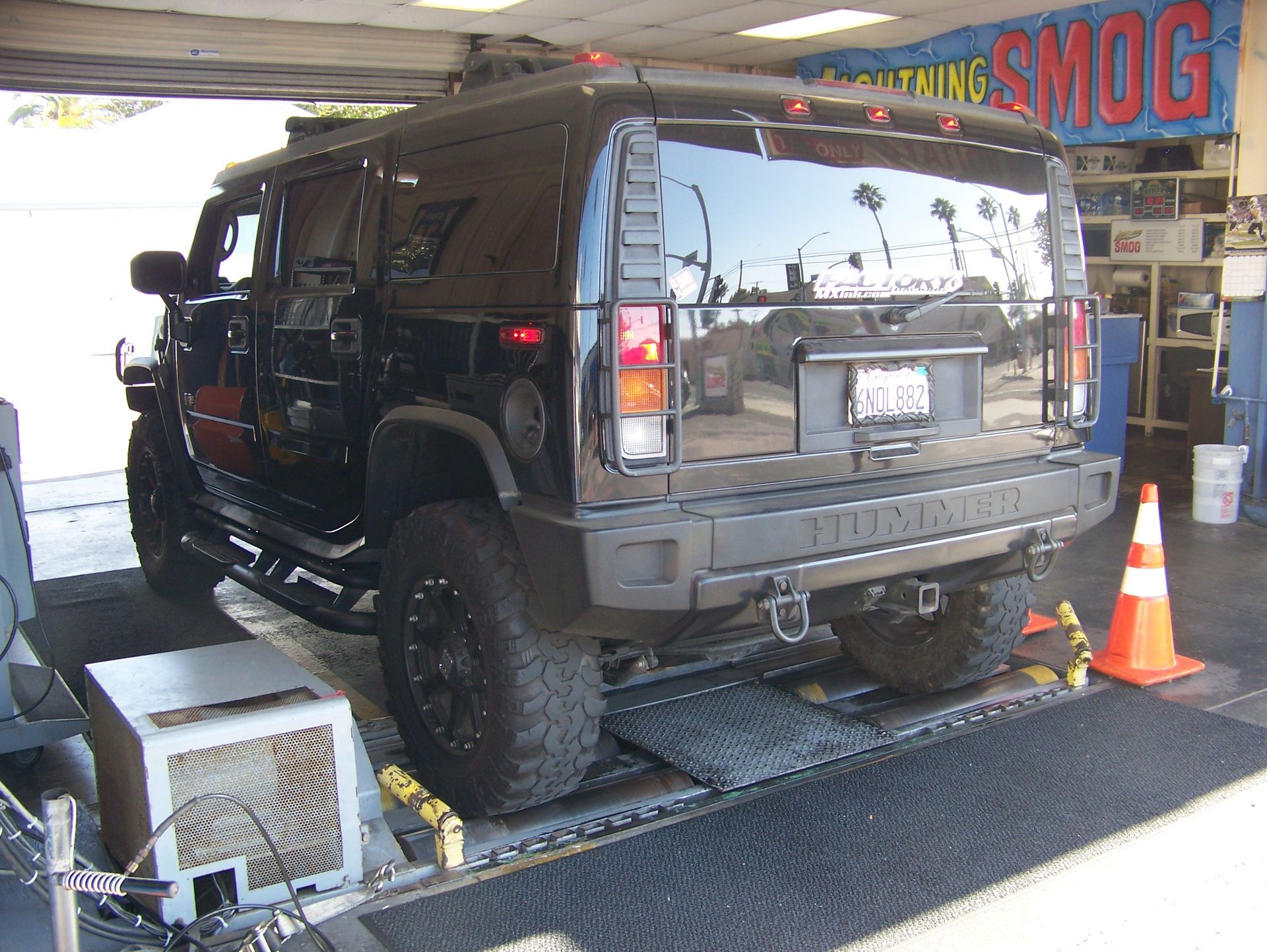 Black SUV on a dynamometer in a smog check garage, with cones and equipment nearby.