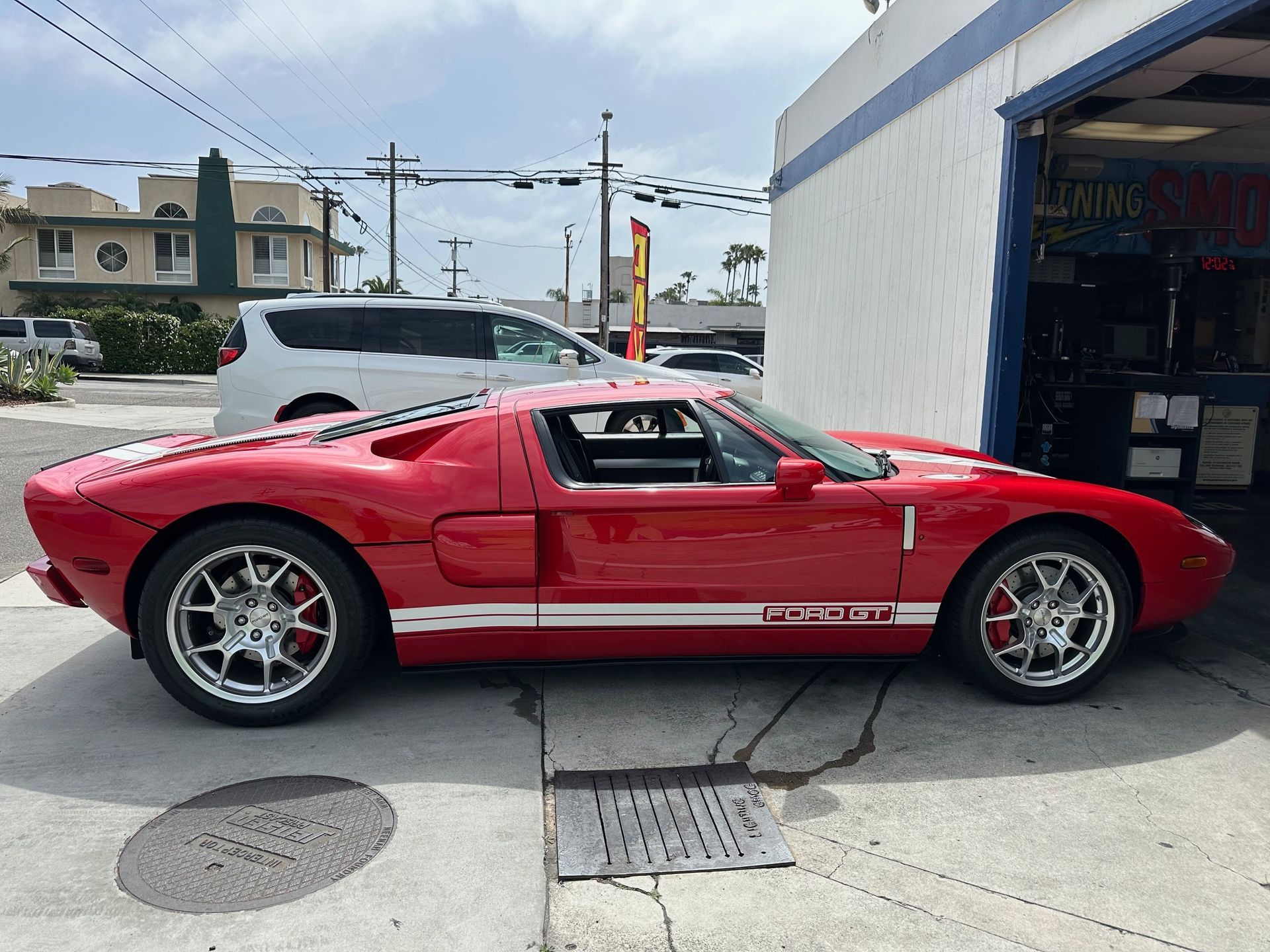 Red sports car parked outside an auto shop, side view with white racing stripes.
