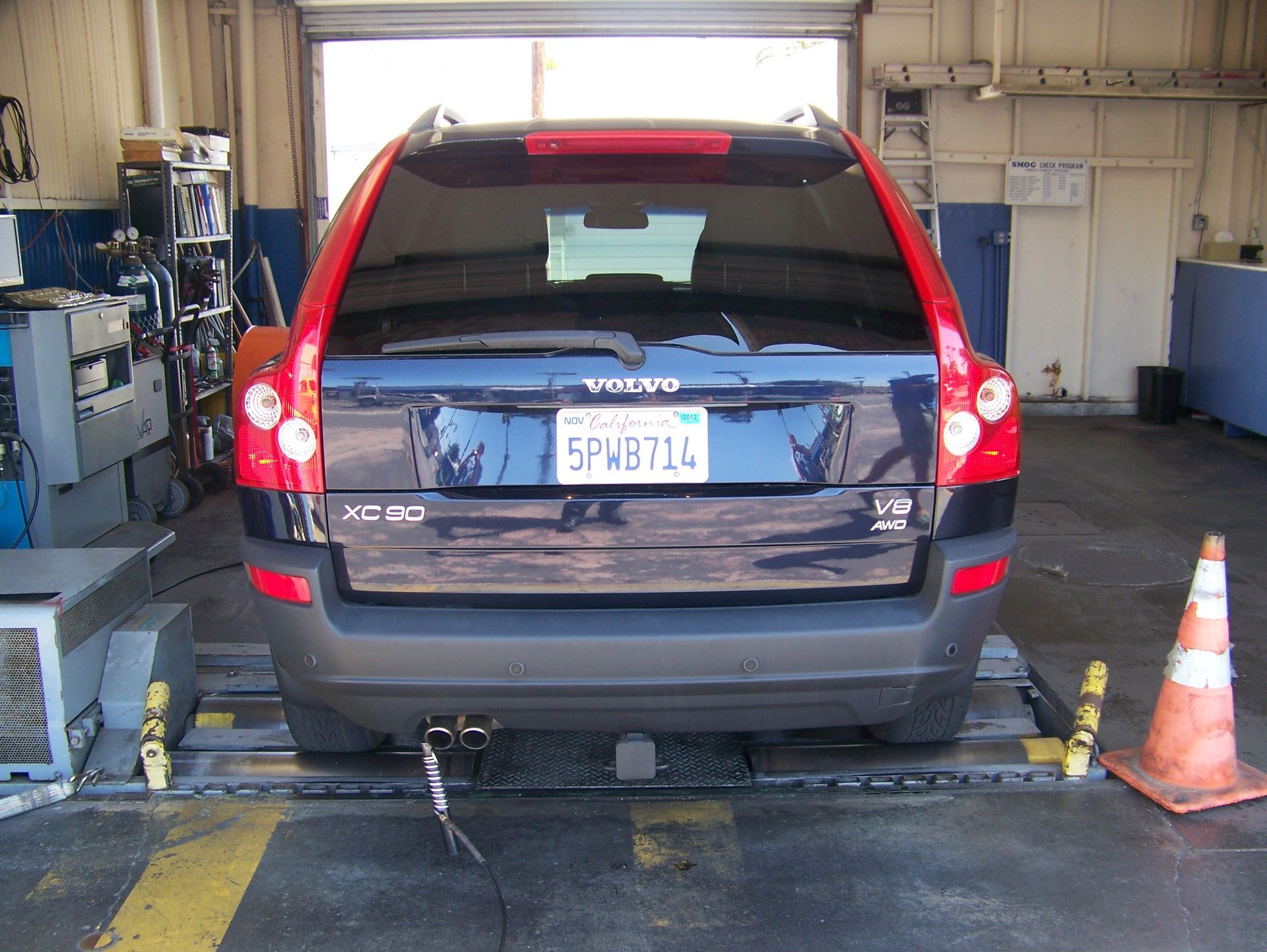 Black SUV on a lift in an auto repair garage, viewed from the rear, with cones and equipment nearby.