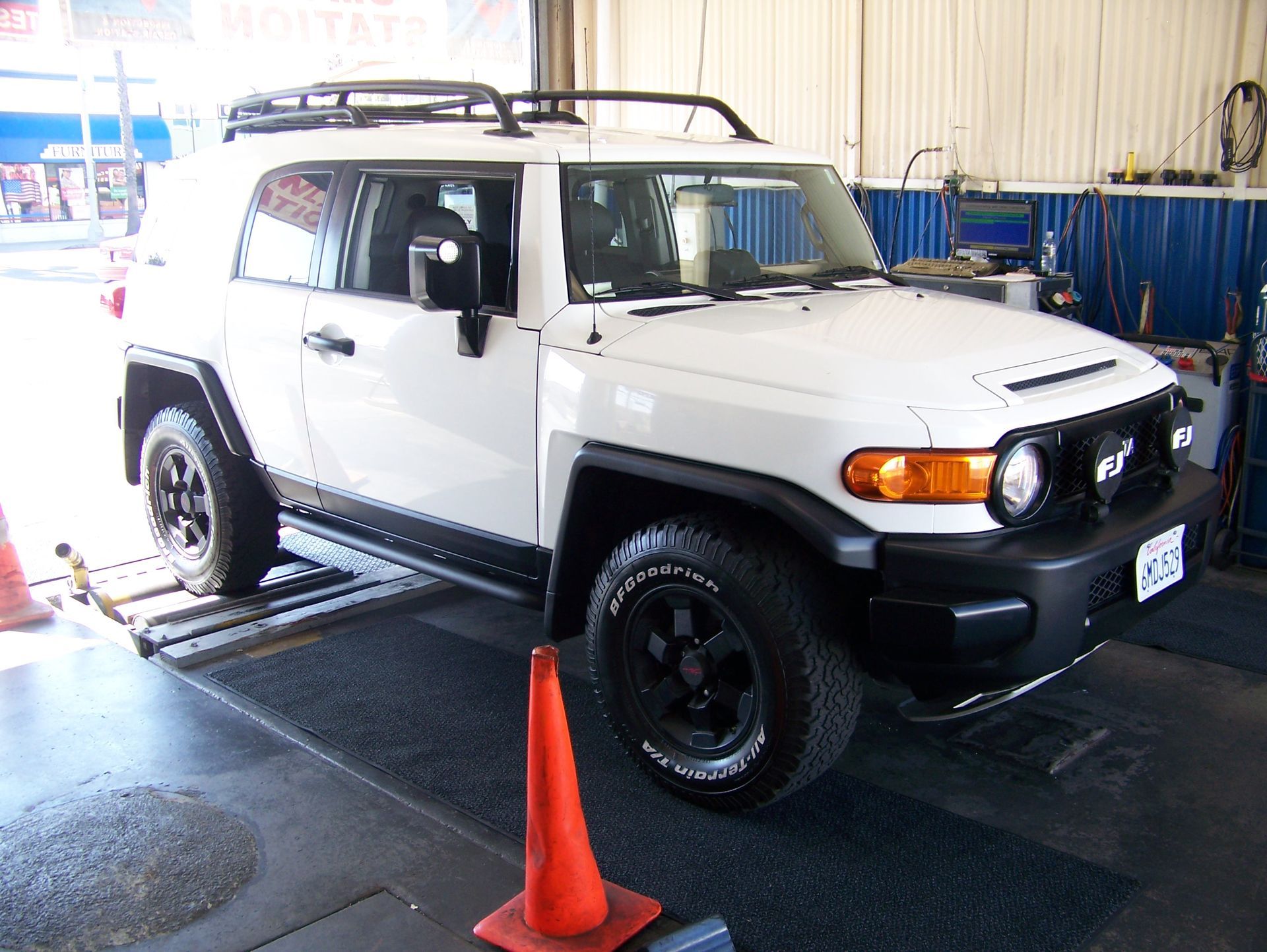 White Toyota FJ Cruiser in an auto shop, with an orange cone in front and black wheels.