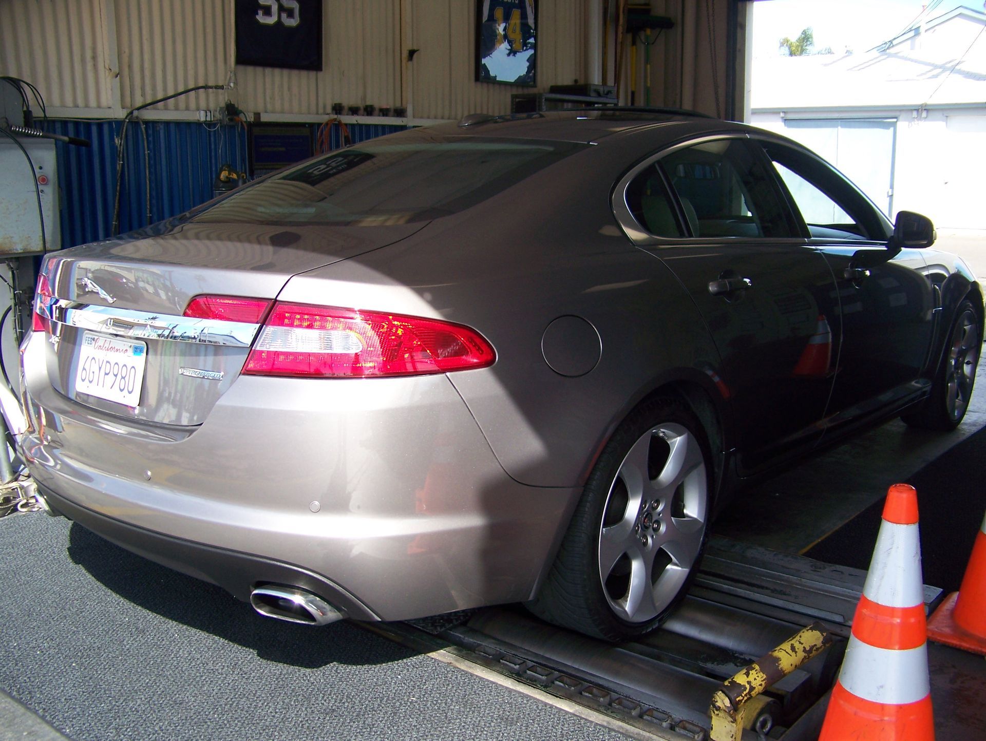 Gray sedan on a dynamometer test stand in a garage, with orange traffic cones nearby.