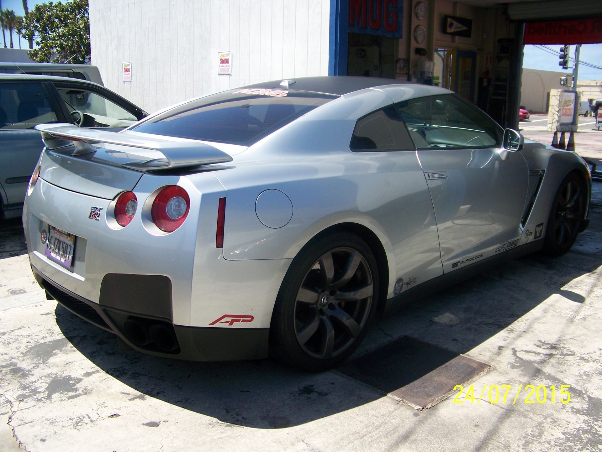 Silver sports car parked on a sunny street, rear three-quarter view, with a black spoiler and tinted windows