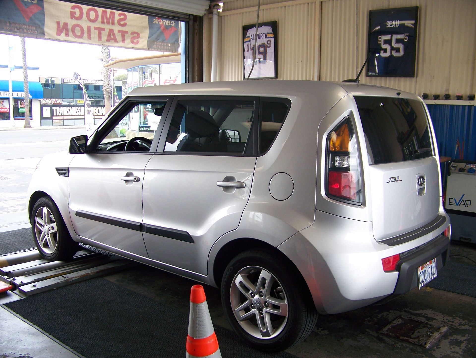 Silver compact car on a dynamometer in a garage, with an orange cone in front.