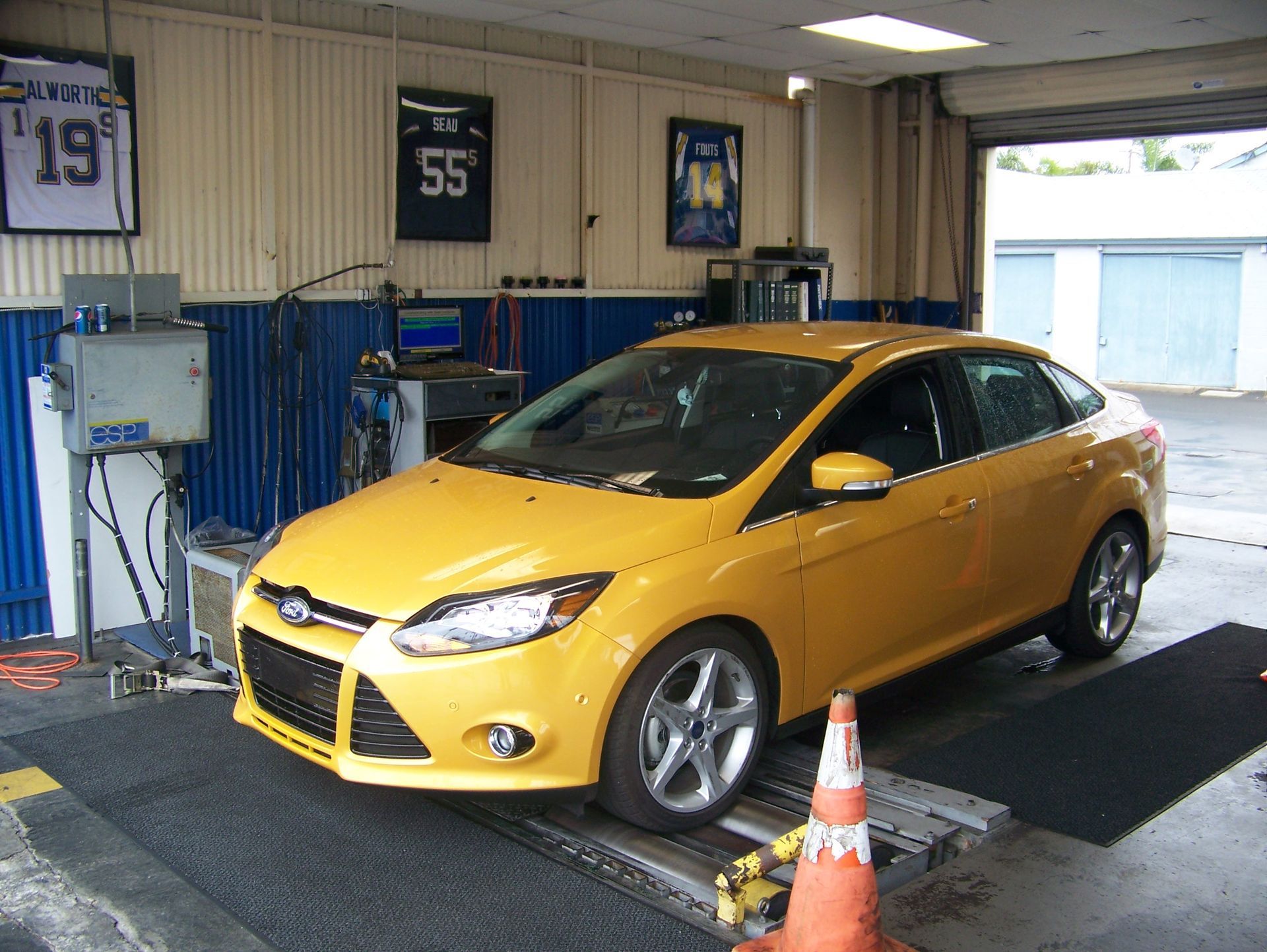 Yellow sedan on a dynamometer in an auto shop garage, with a traffic cone in front