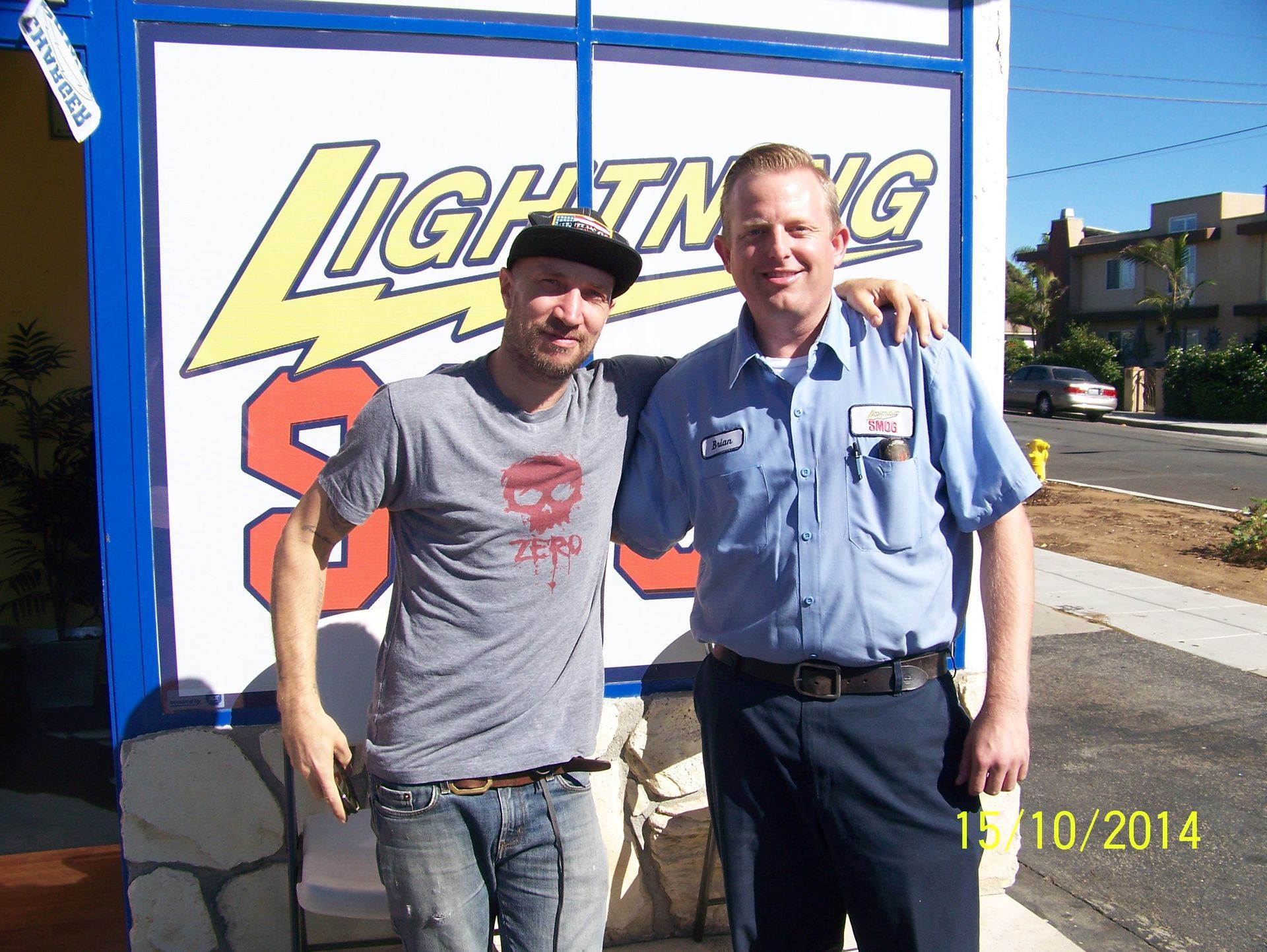 Two smiling men pose in front of a Lightning sign outdoors.
