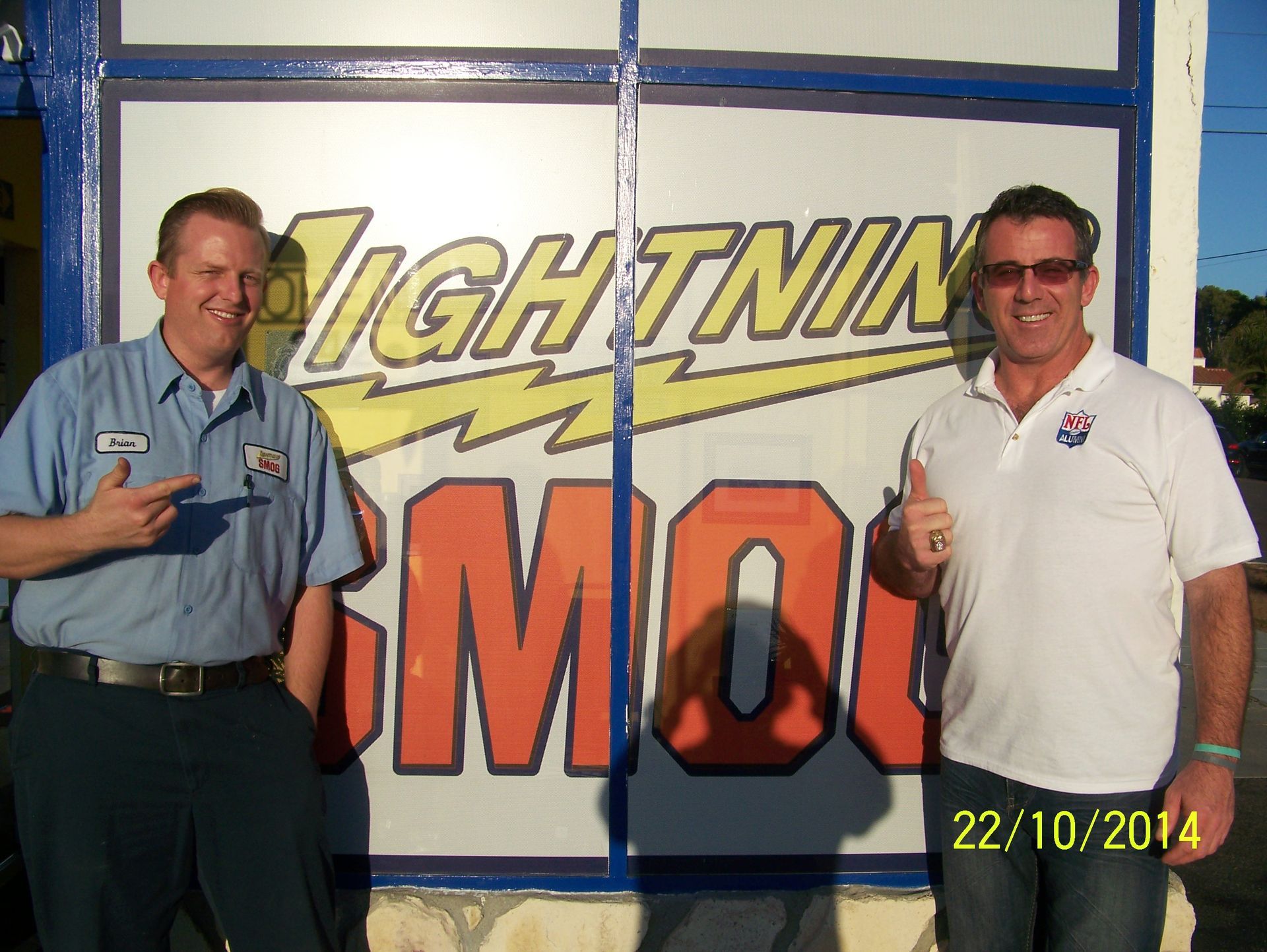 Two men posing in front of a Lightning Armor sign, both holding up keys and smiling.