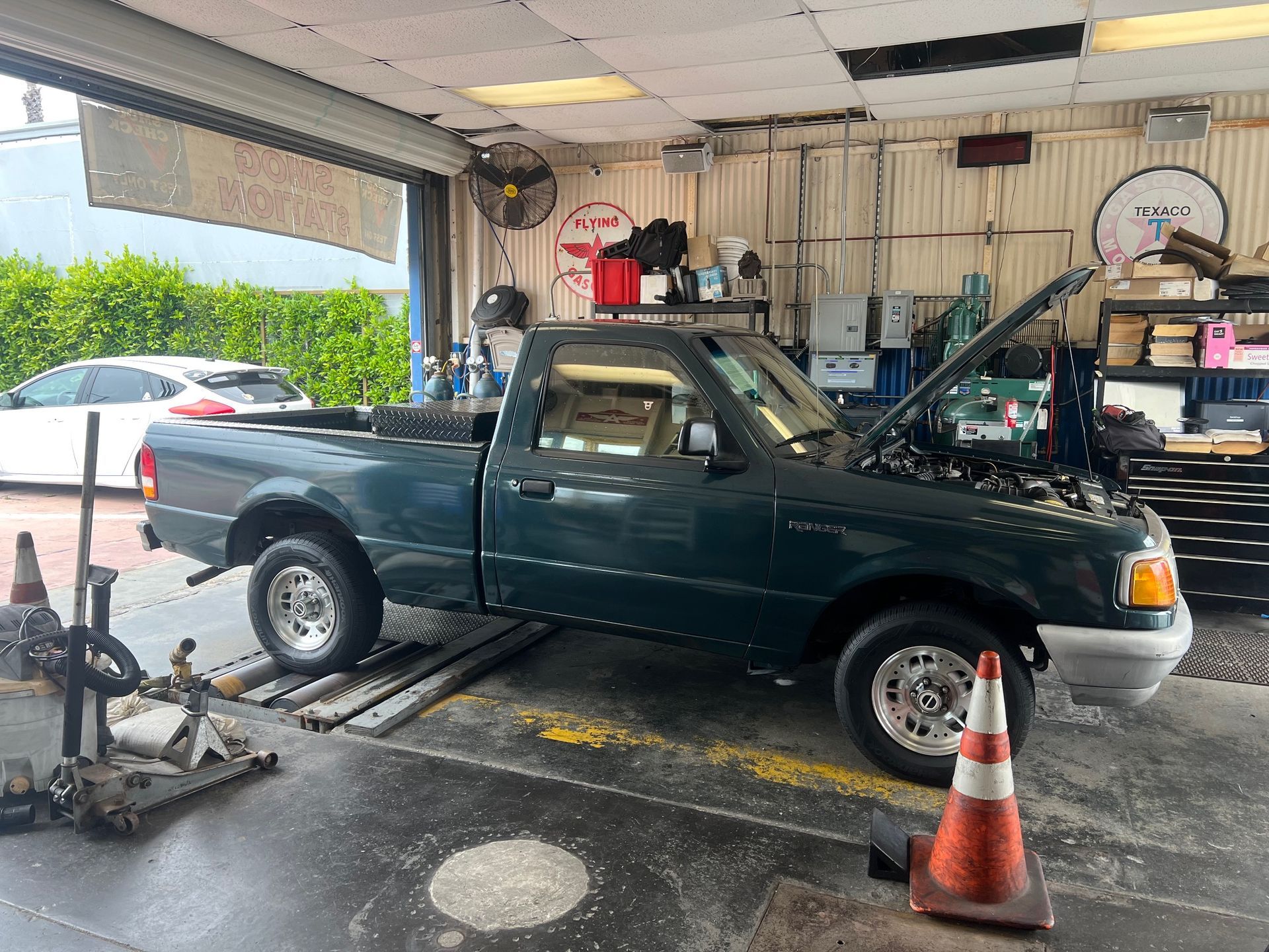 Dark green pickup truck on a repair shop lift with a raised hood and an orange cone nearby