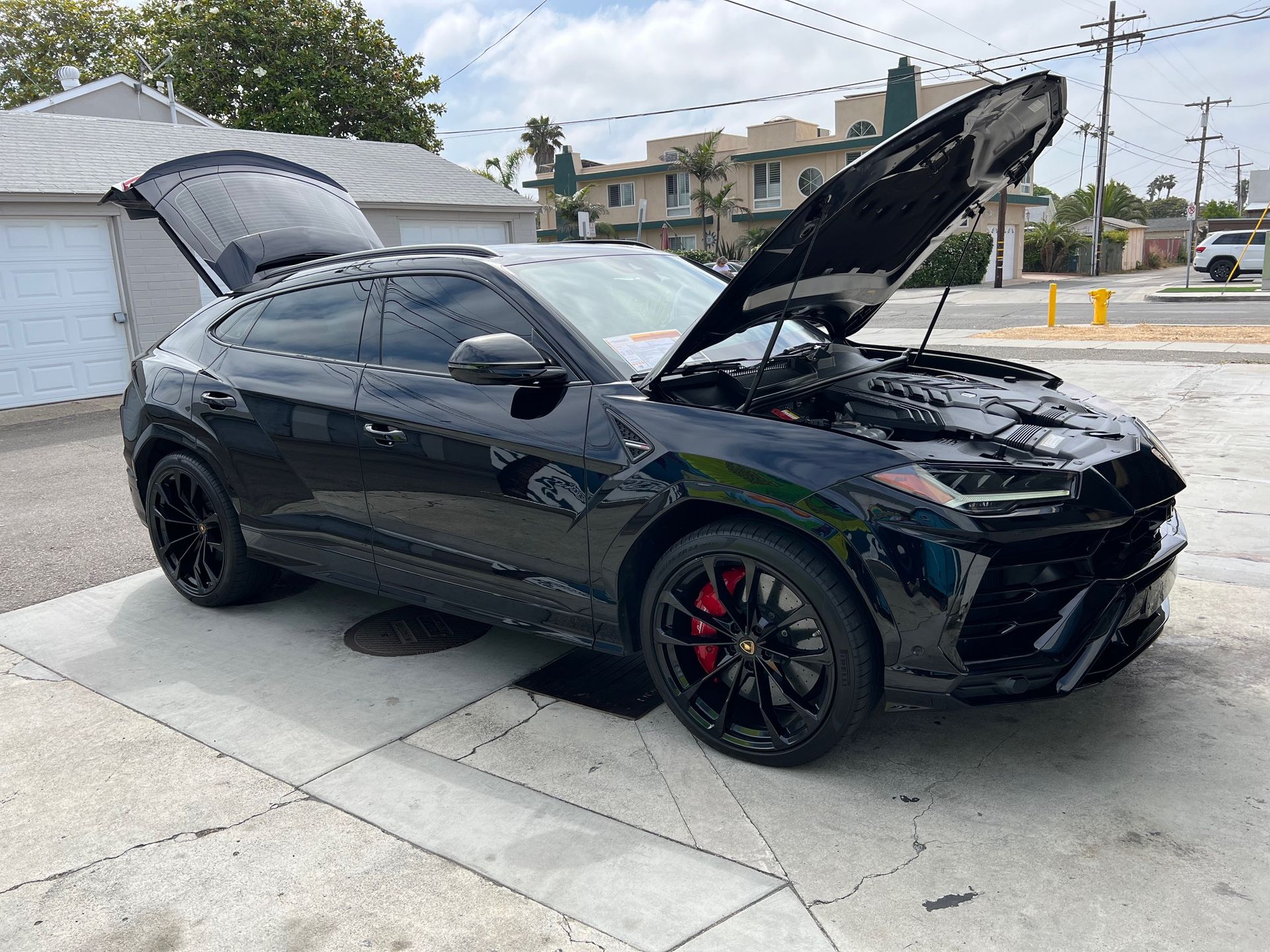 Black sports coupe with hood open in a driveway, showing red brake calipers and black wheels.