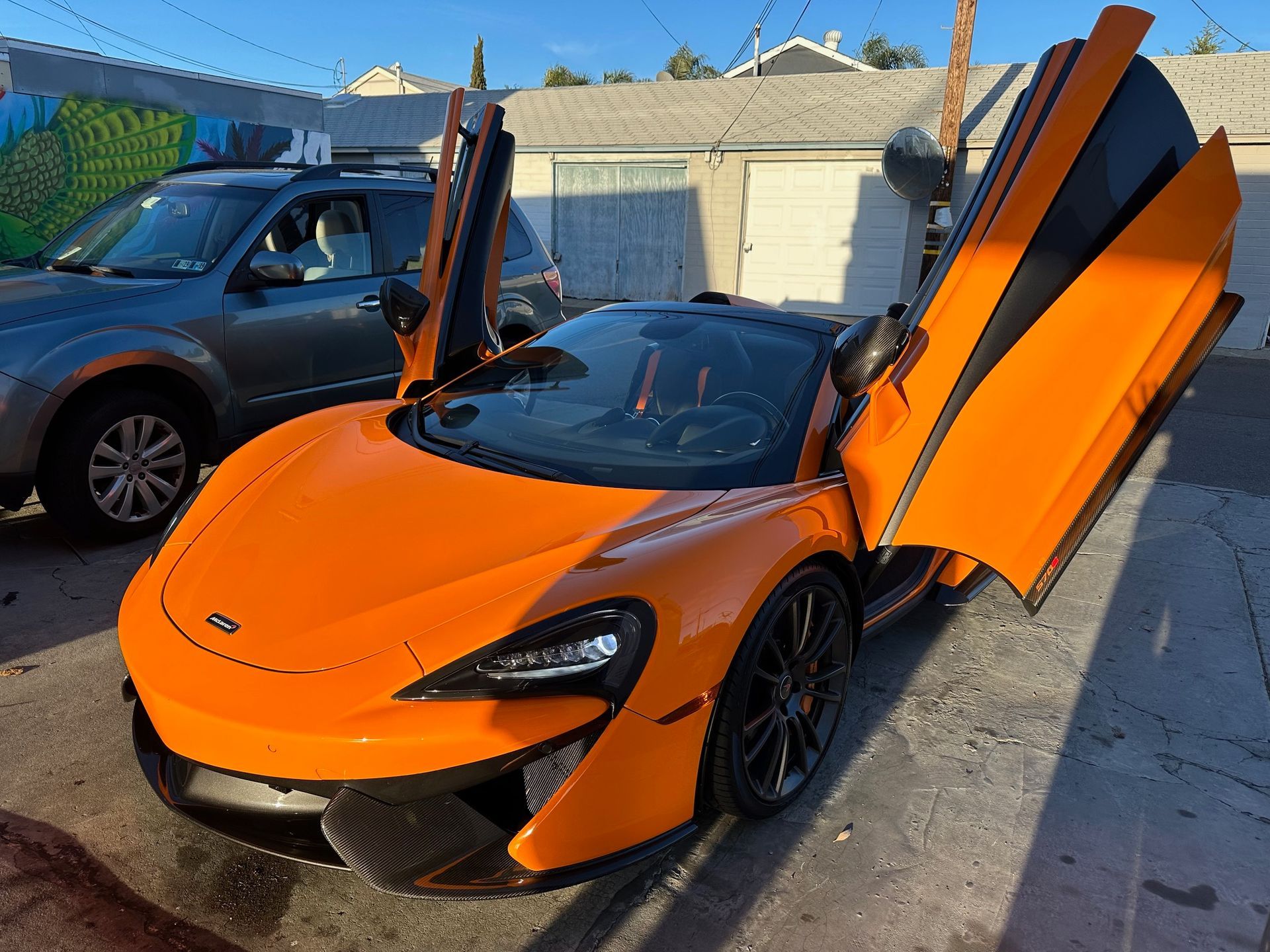 Bright orange sports car with scissor doors open in a driveway, beside a gray SUV.