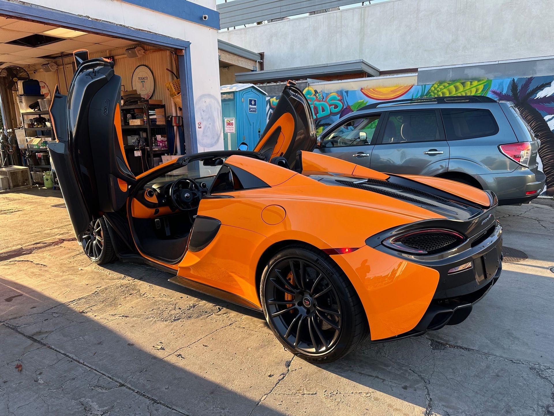 Orange sports car with scissor door open in a garage, with a blue SUV parked behind it.