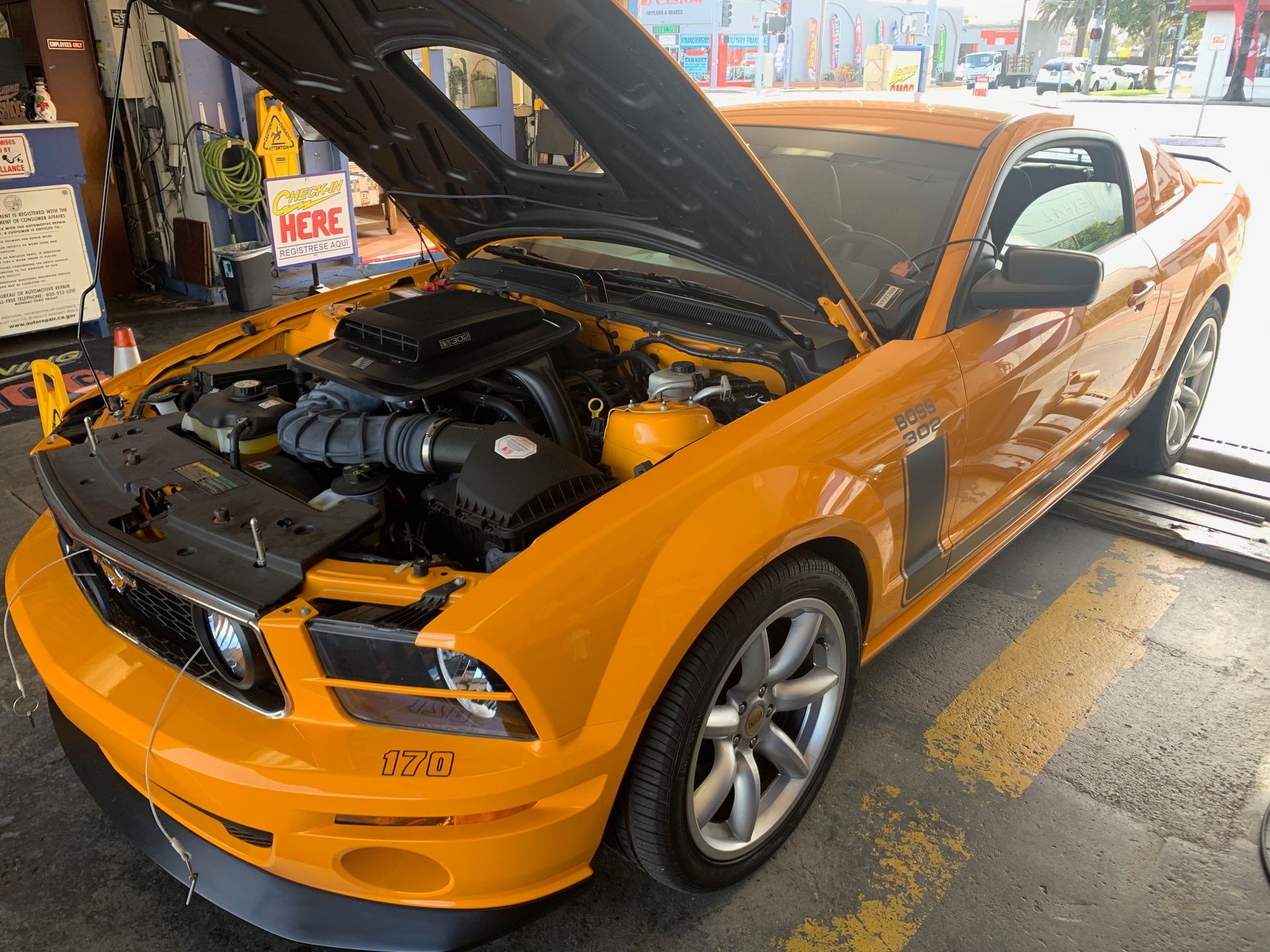 Orange Ford Mustang with hood open in a garage, showing the engine bay.