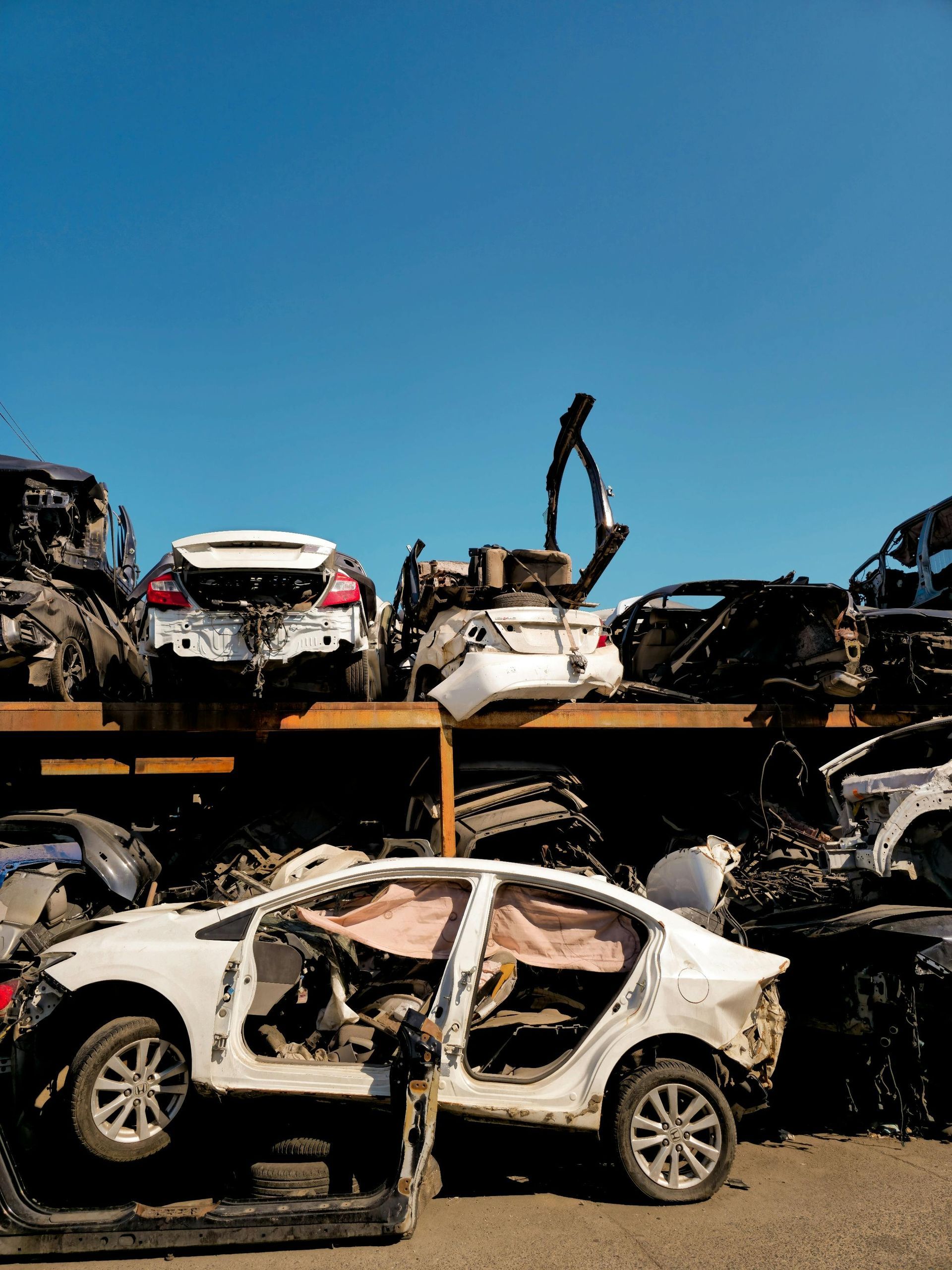 White cars stacked in a scrapyard under a bright blue sky, with a crane arm above.