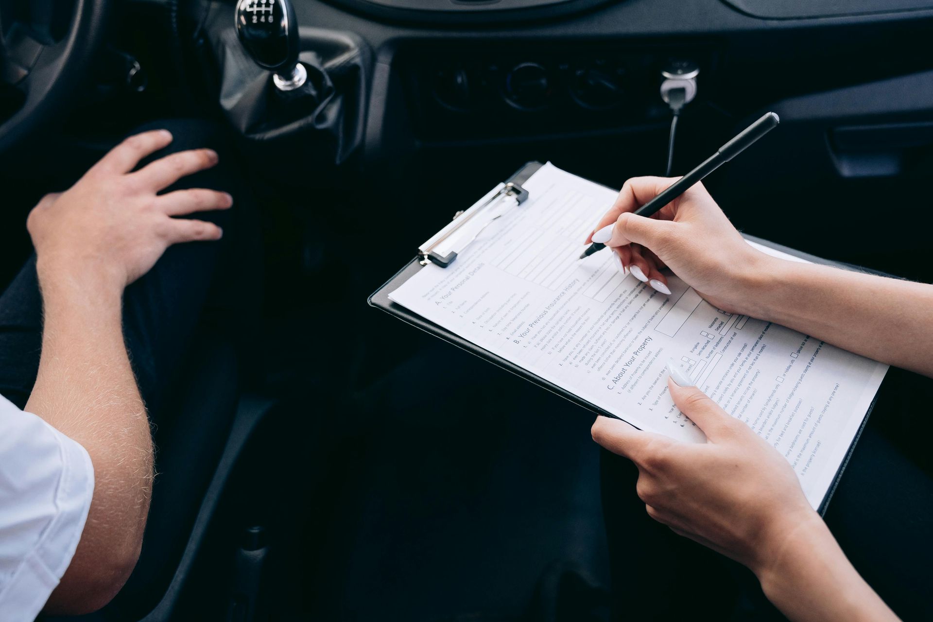 Hands writing on a clipboard beside a car steering wheel and dashboard in the dark interior