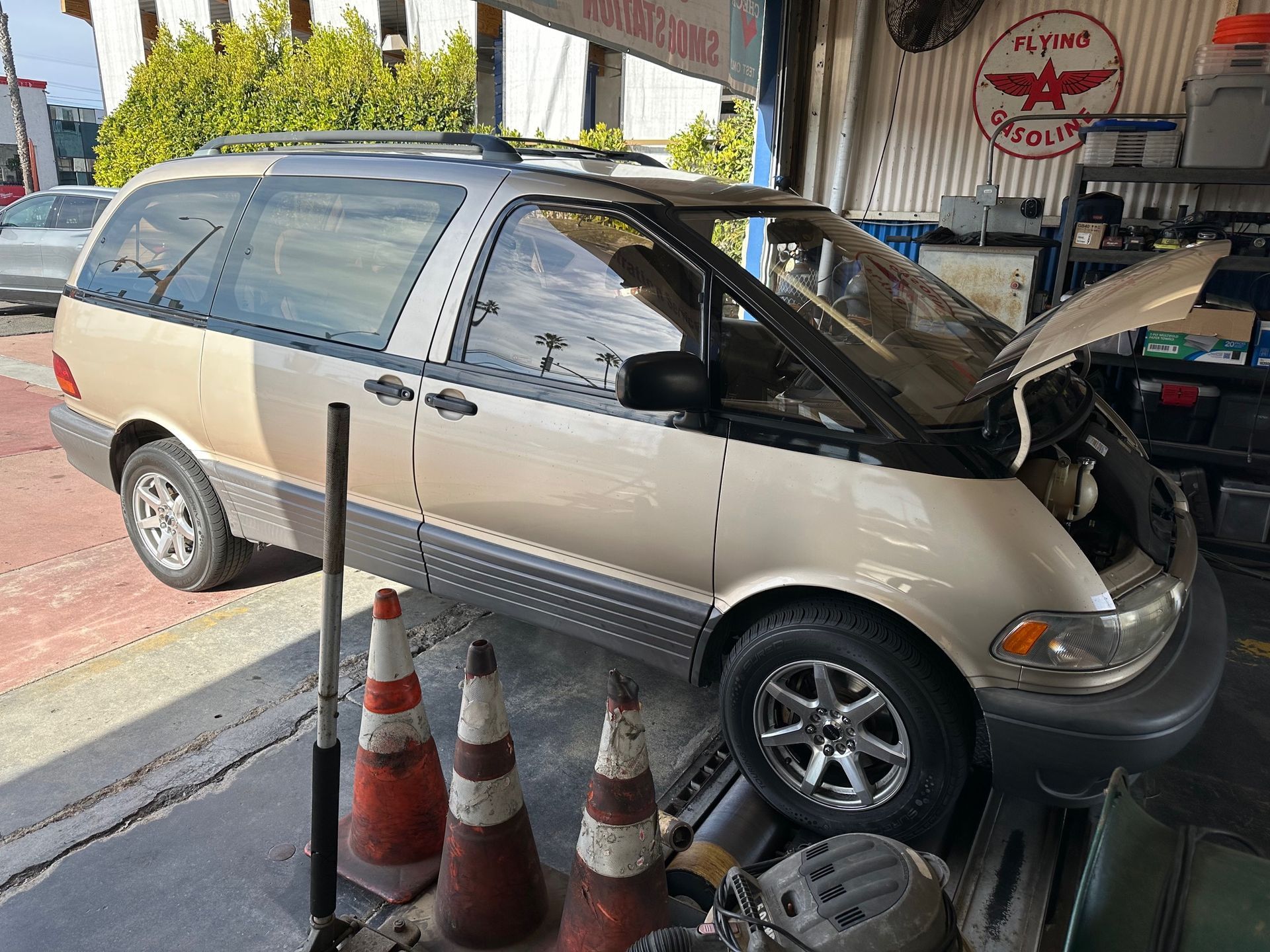 Minivan with hood open in a shop bay, parked beside traffic cones under a covered service area.
