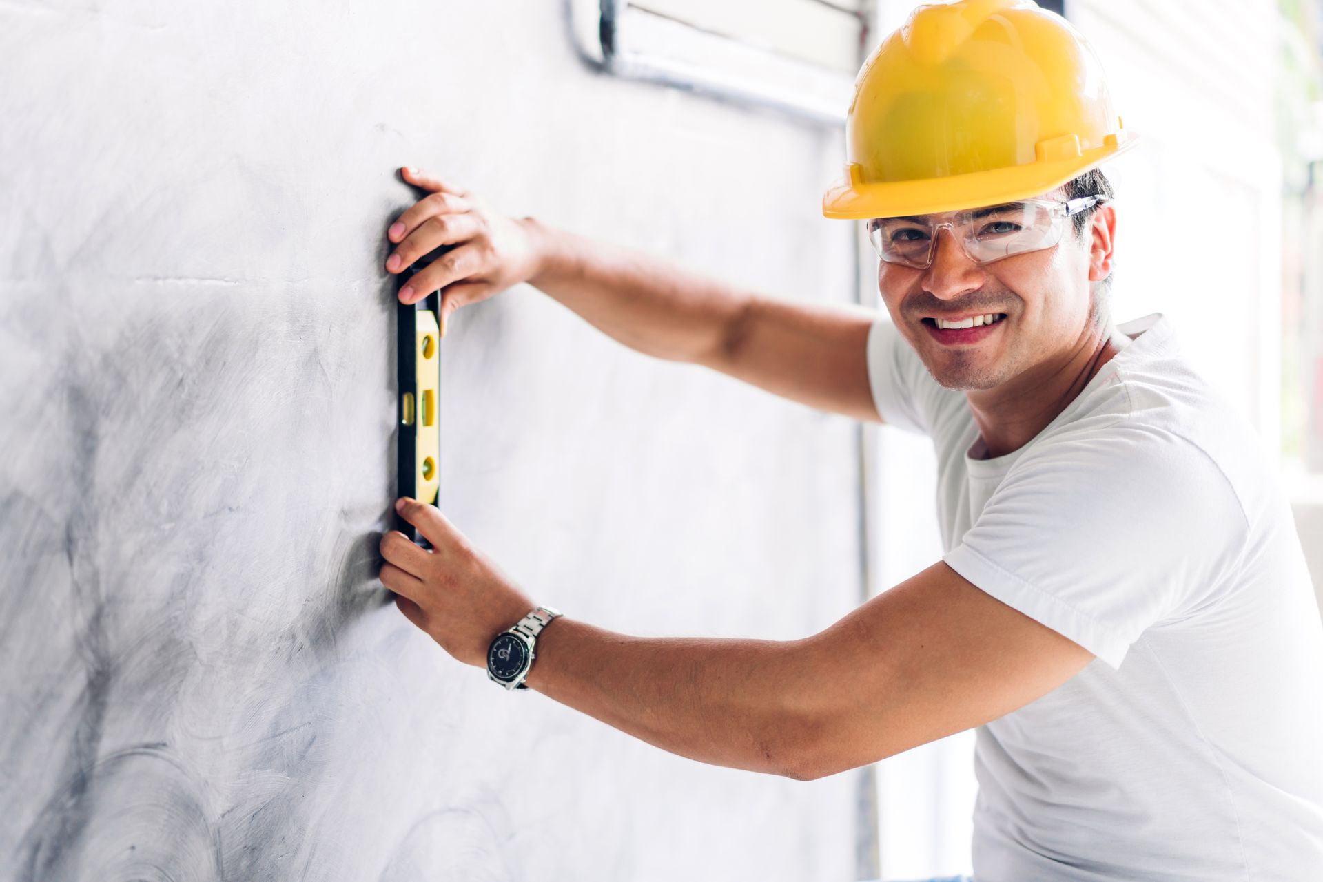 a man wearing a hard hat and safety glasses is measuring a wall with a level .
