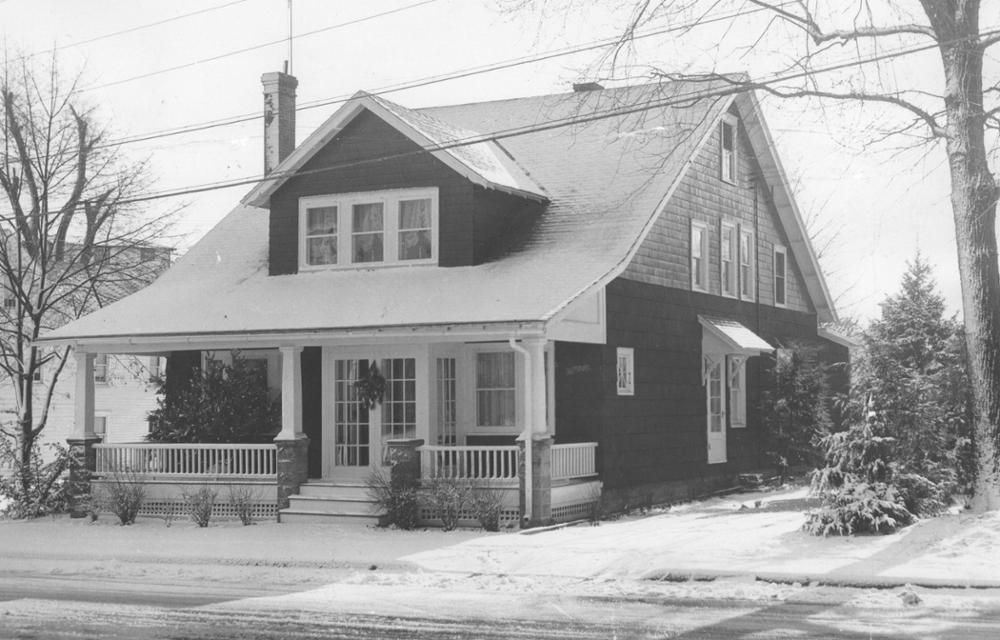 Snow-covered house with a porch and bare trees in winter.